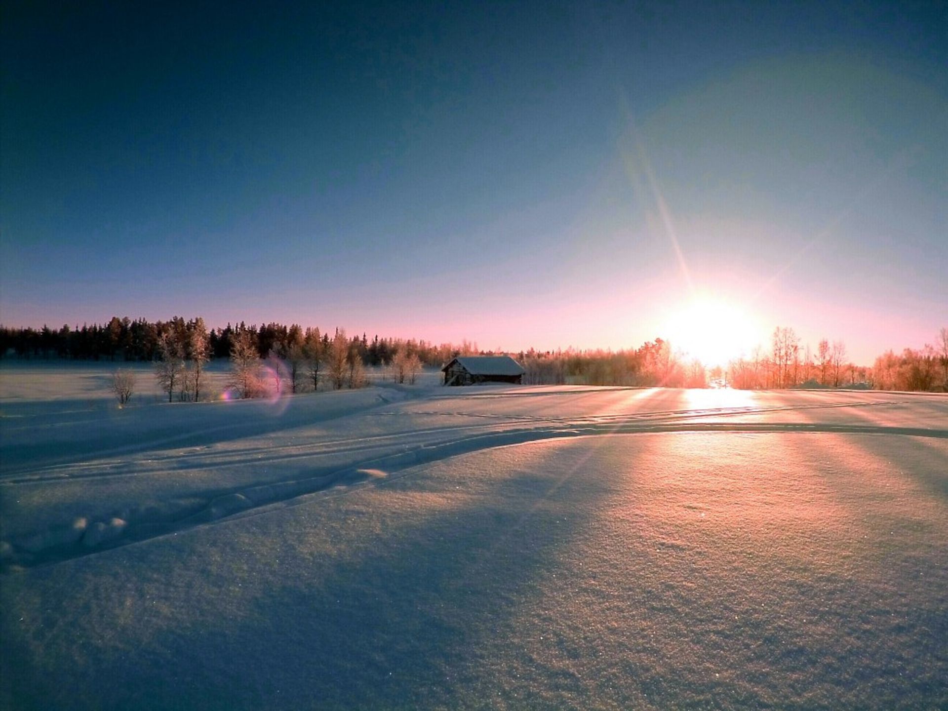 A landscape where the white snow is visible and the sun is on its way down behind the green forest.