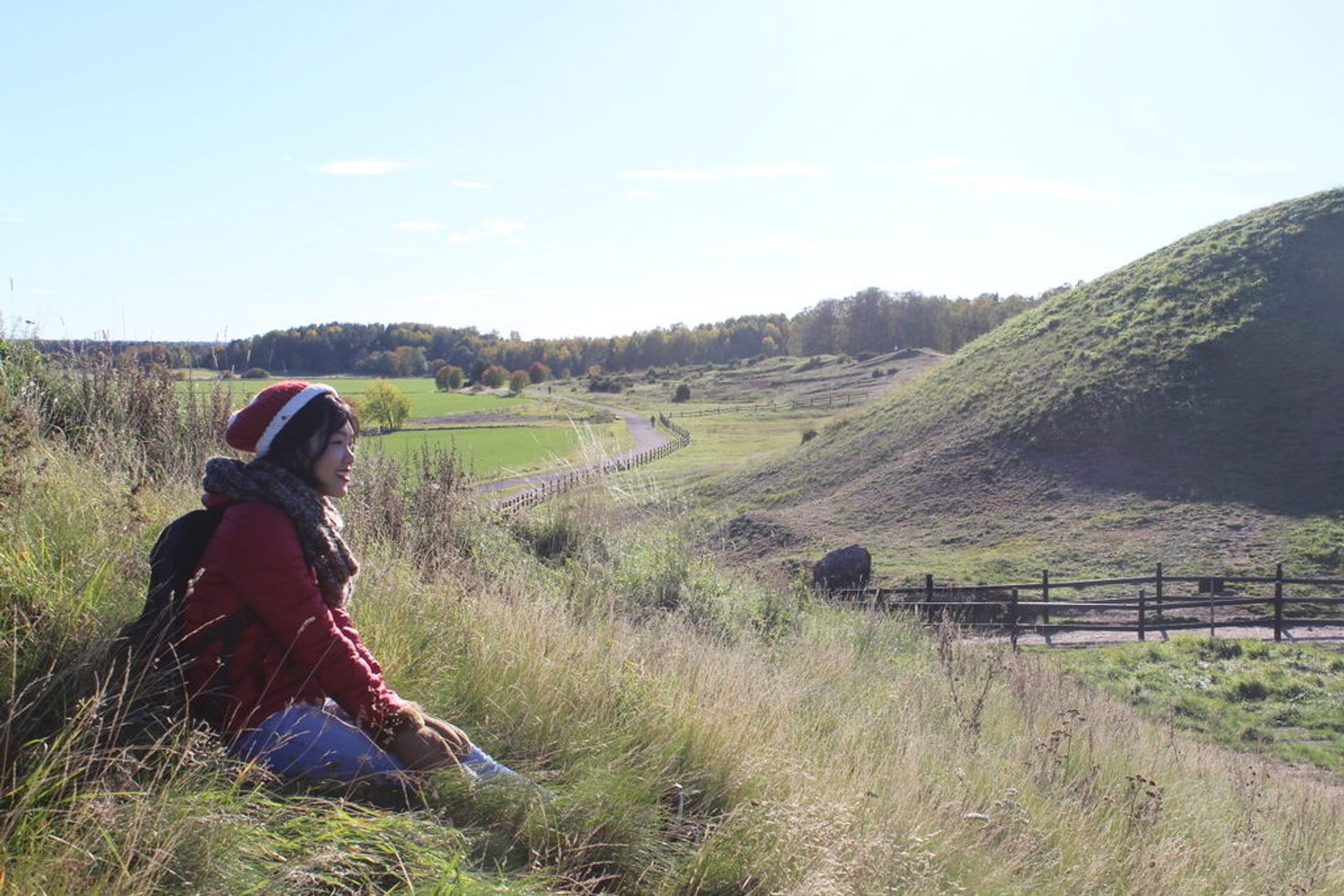 A woman sitting on a hill full of long grease. In the background, it is another hill and a wooden railing.