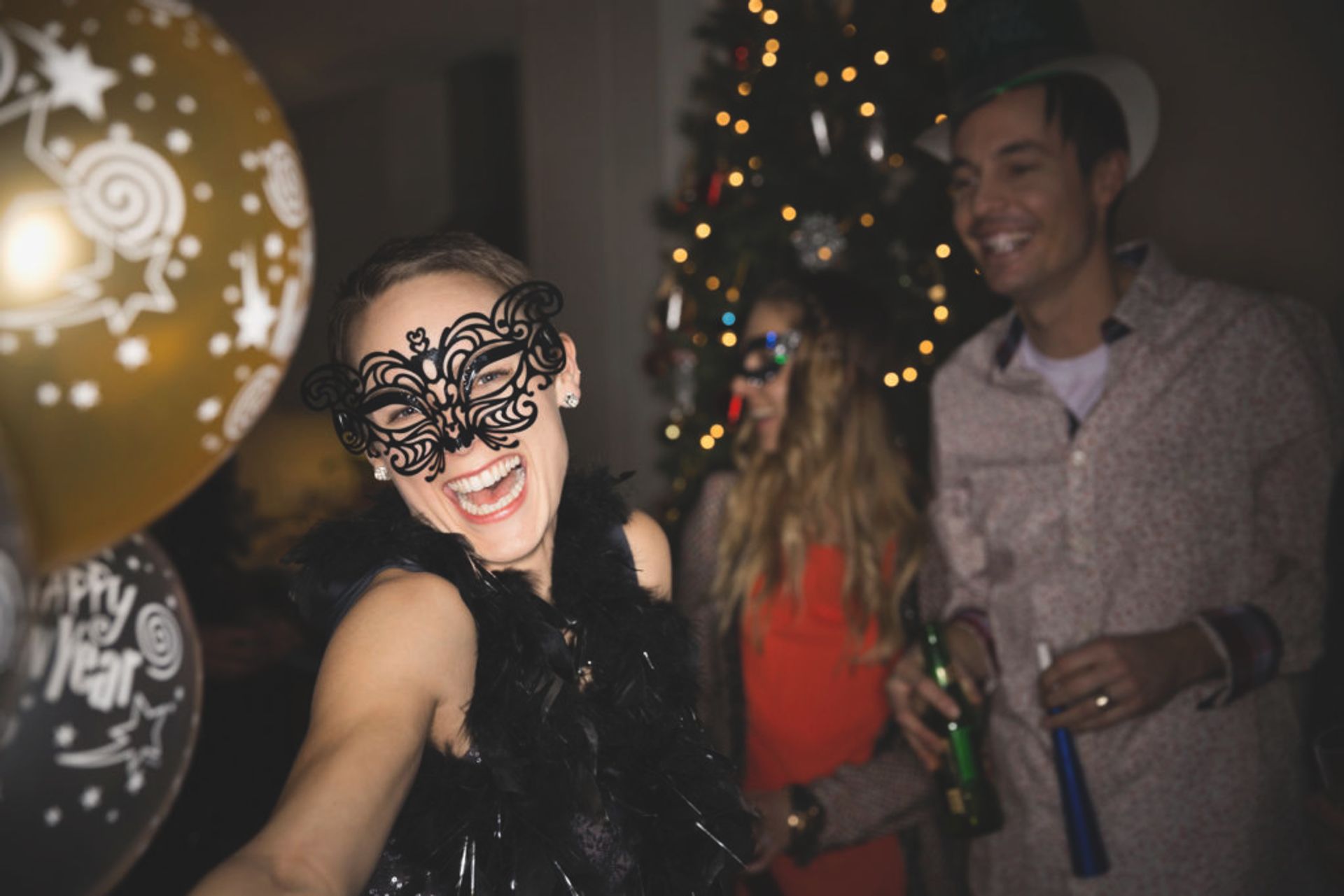 People having a great time at a party. A woman is smiling at the camera with a black decorated mask. It is a man and a woman smiling in the background.
