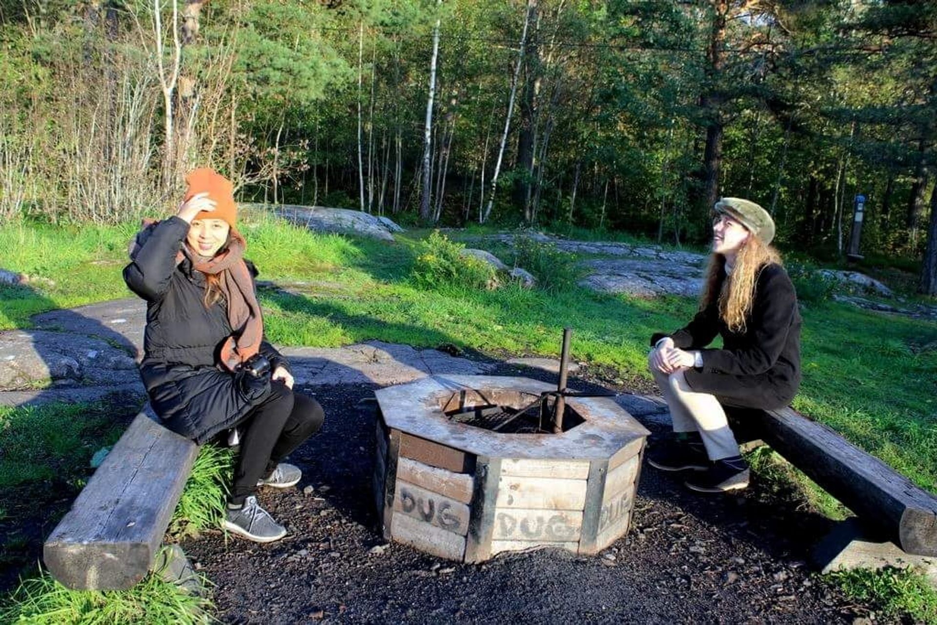Two women by a fireplace in the forest. It is chilly as both are wearing winter jackets.
