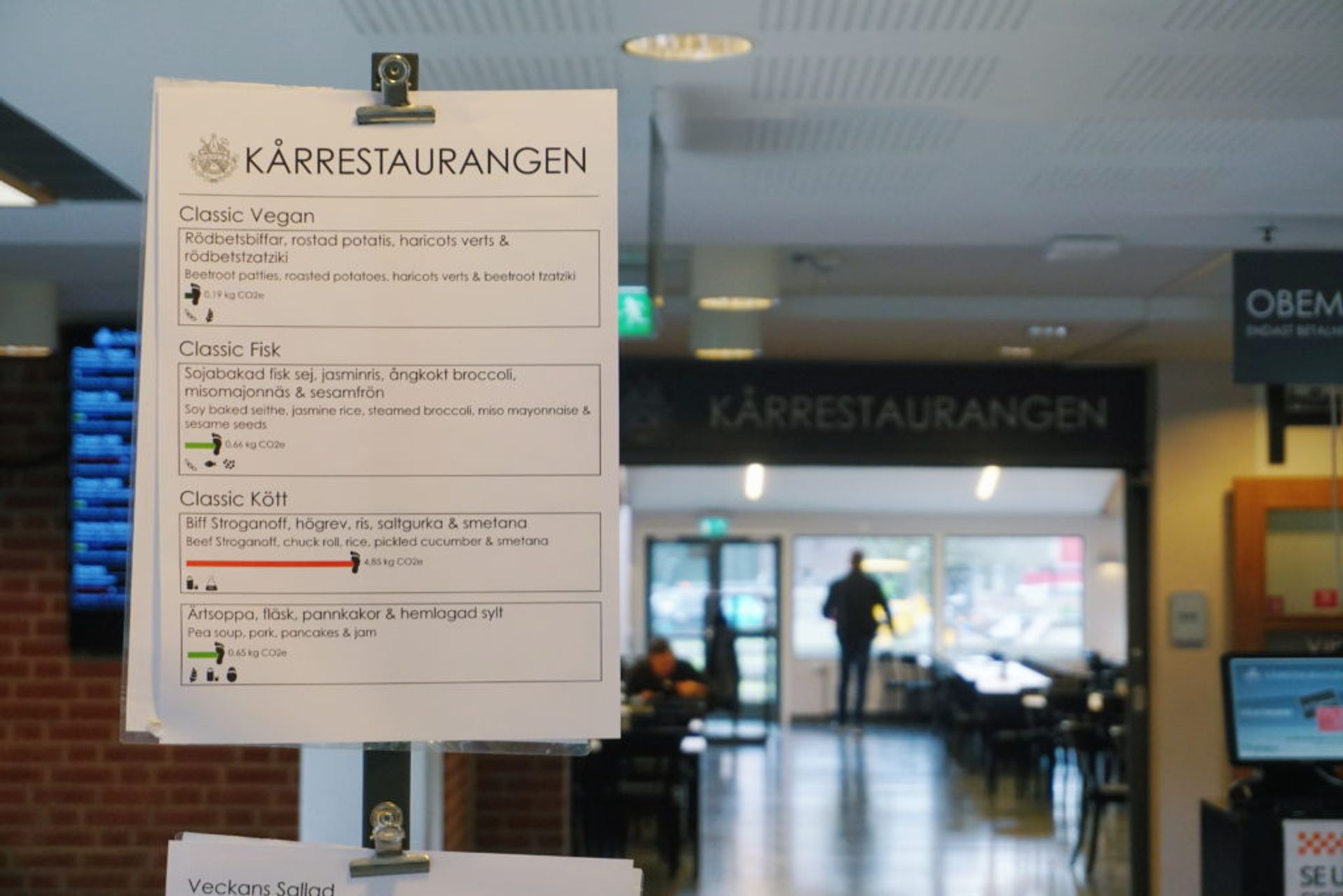A menu with vegan, fish and meat option, from the restaurant "Kårrestaurangen", which is seen in the background.