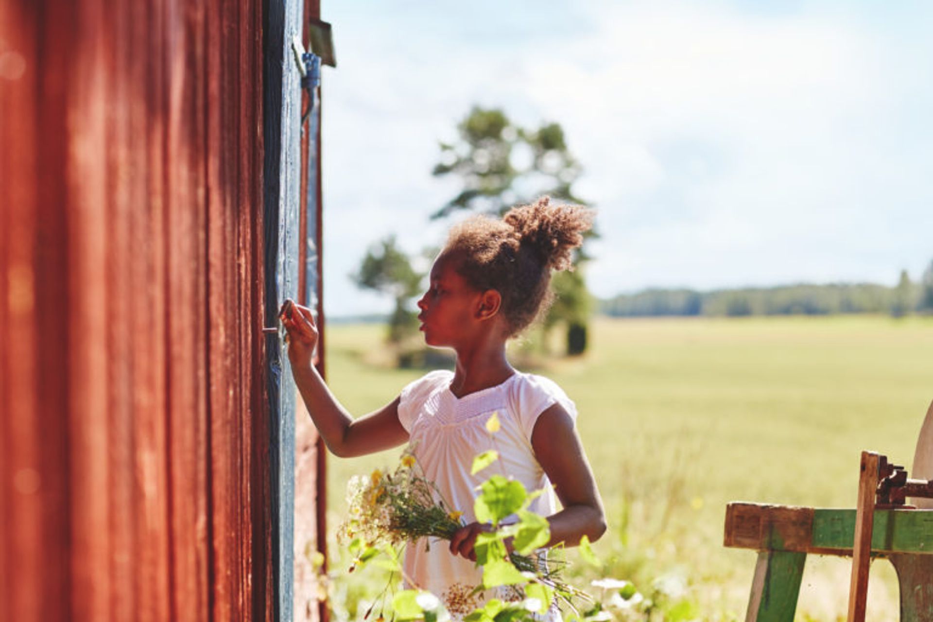 A child is crafting by a red house, right by a meadow, on a sunny summer day. 