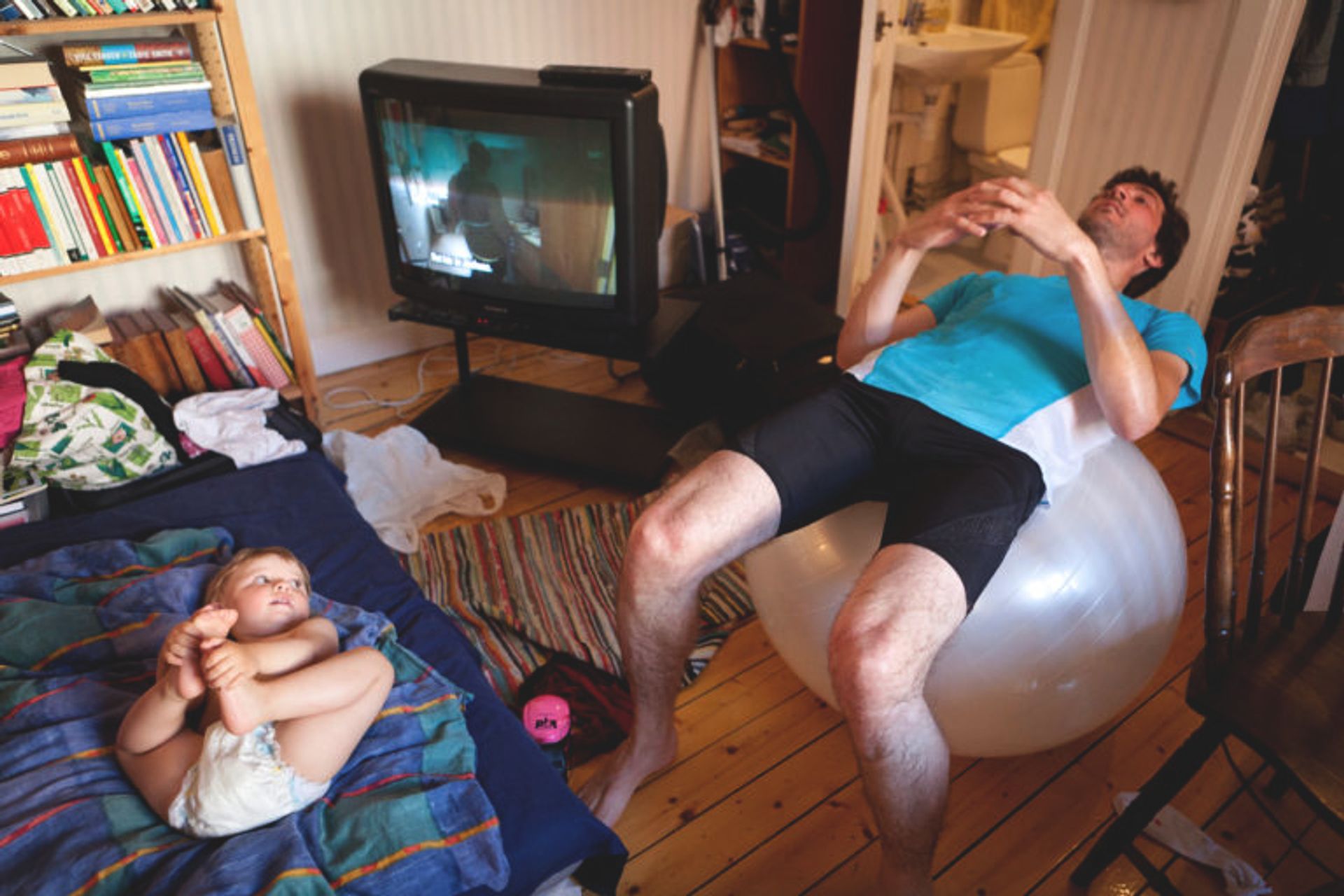 A father is working out at home with a pilates ball while the baby lies on the bed right next to him.