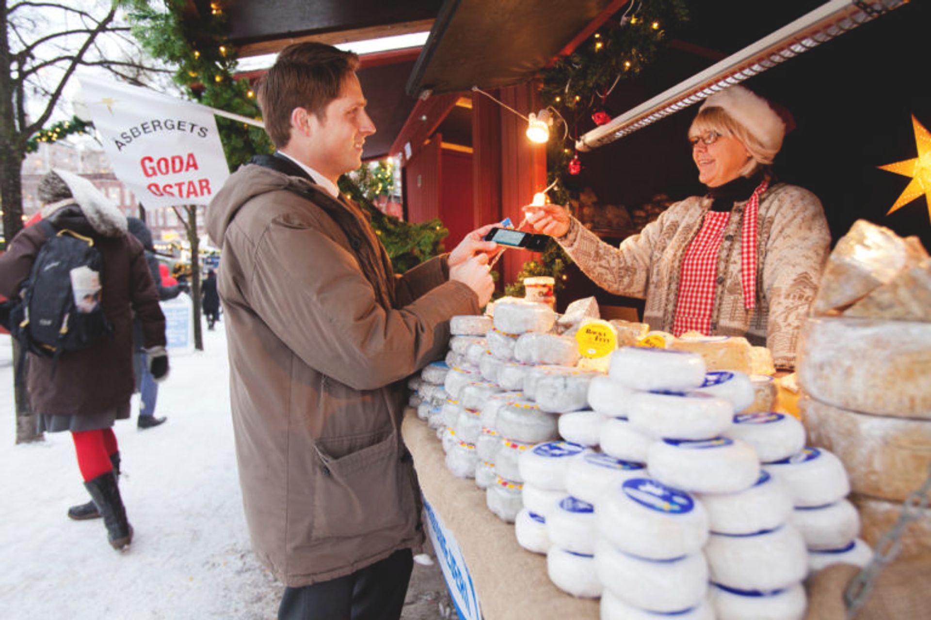 A man buying cheese at a cheese stand from the woman working. It is a cloudy day and it is at a Christmas market. 