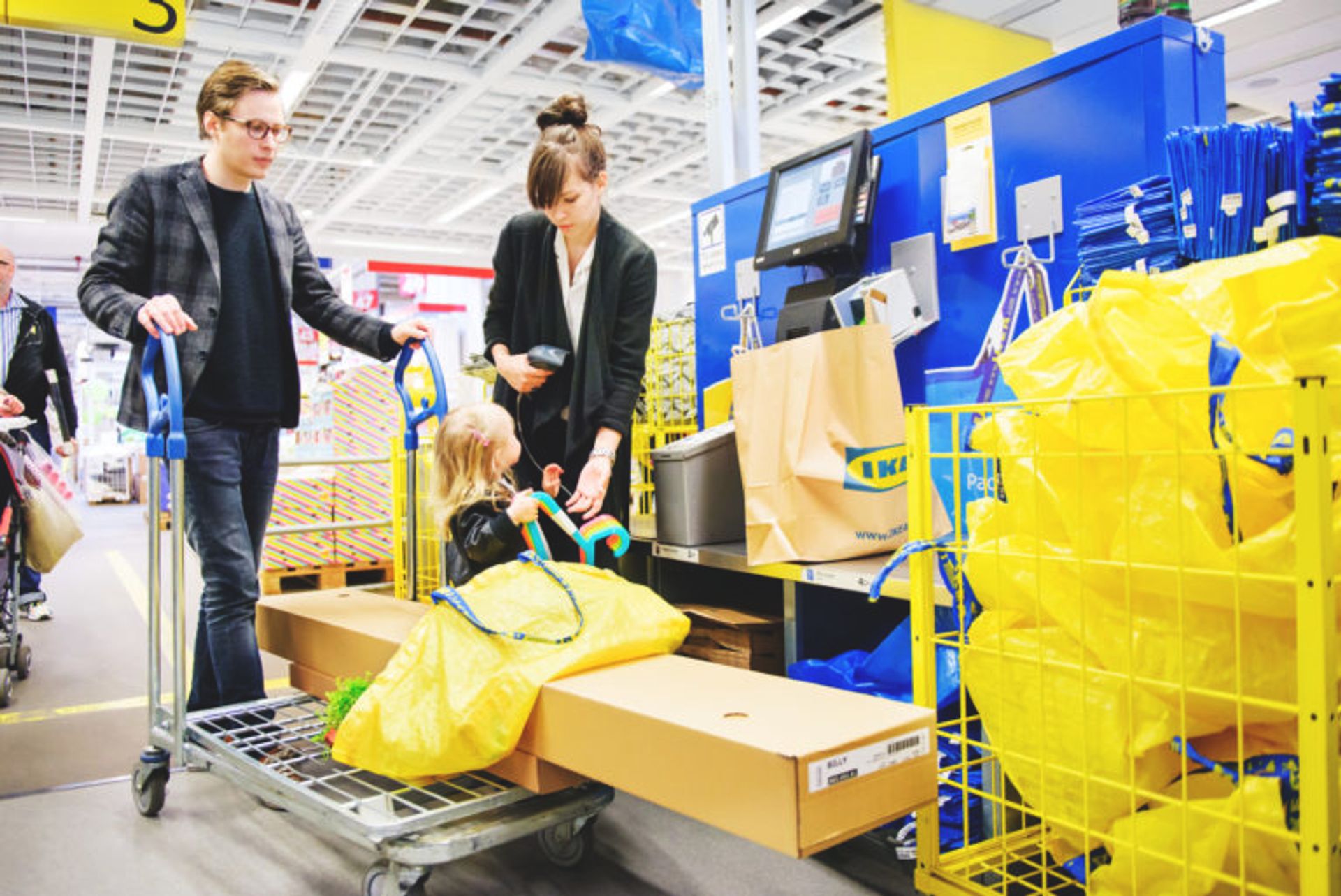 A family at the self check-out at IKEA, scanning all their items. The child is sittning upon the shopping chart, the mum is scanning and the dad is behind the chart.  