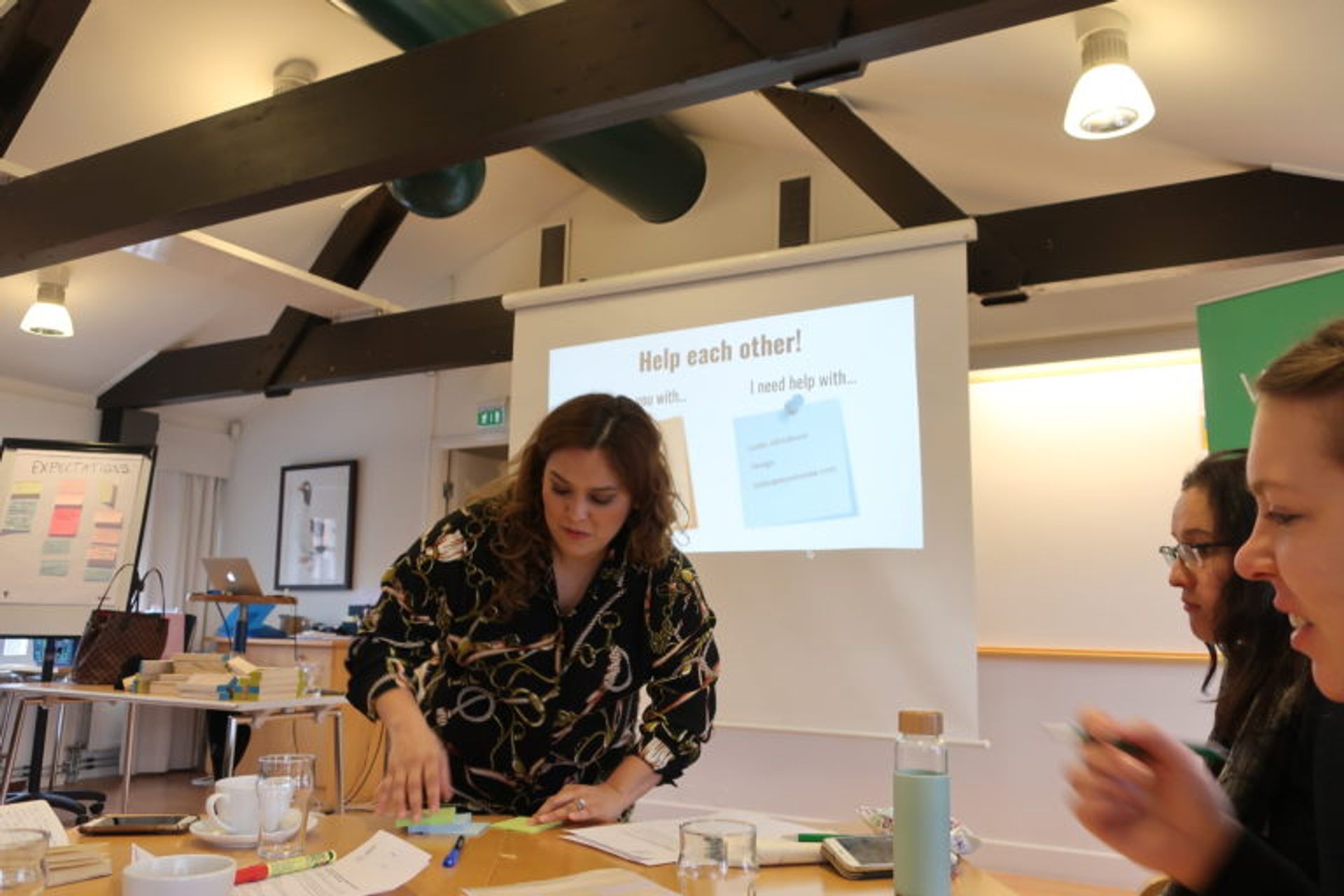 An ongoing workshop where a woman is about to write on a post-it while two other women are looking at her. In the background, you can see a white big screen saying "help each other".