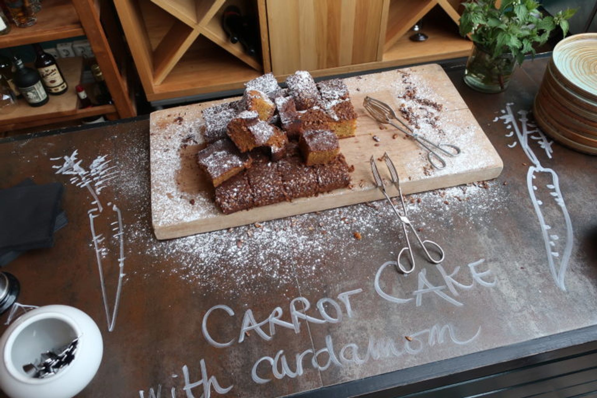 Fika bread on a brown table. It is written "Carrot cake with cardamon" and two carrots, with a white pencil on the table.