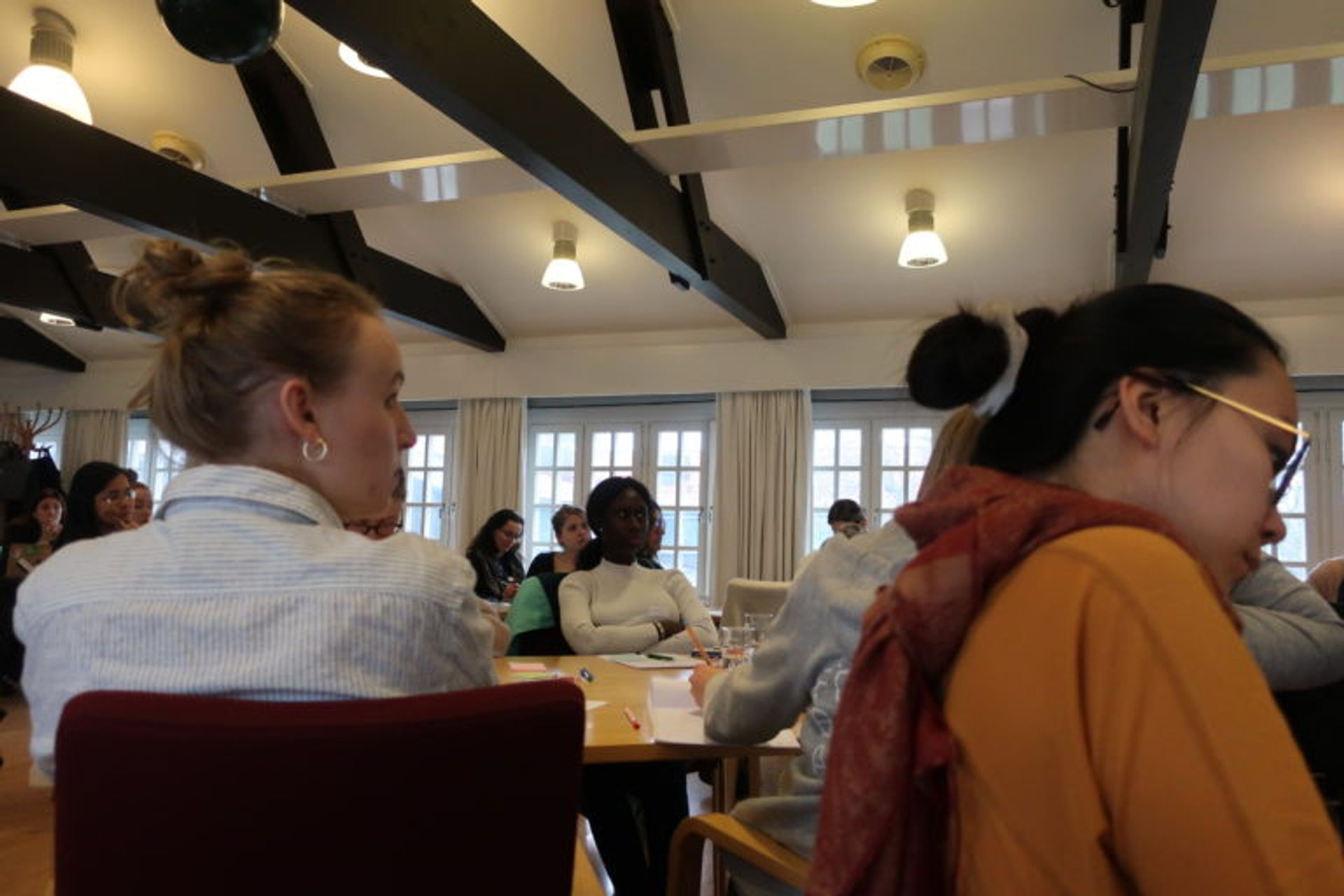 A group of women in a big room with a high ceiling with wooden beams. They are all focused at the front of the room as if someone is standing there talking.