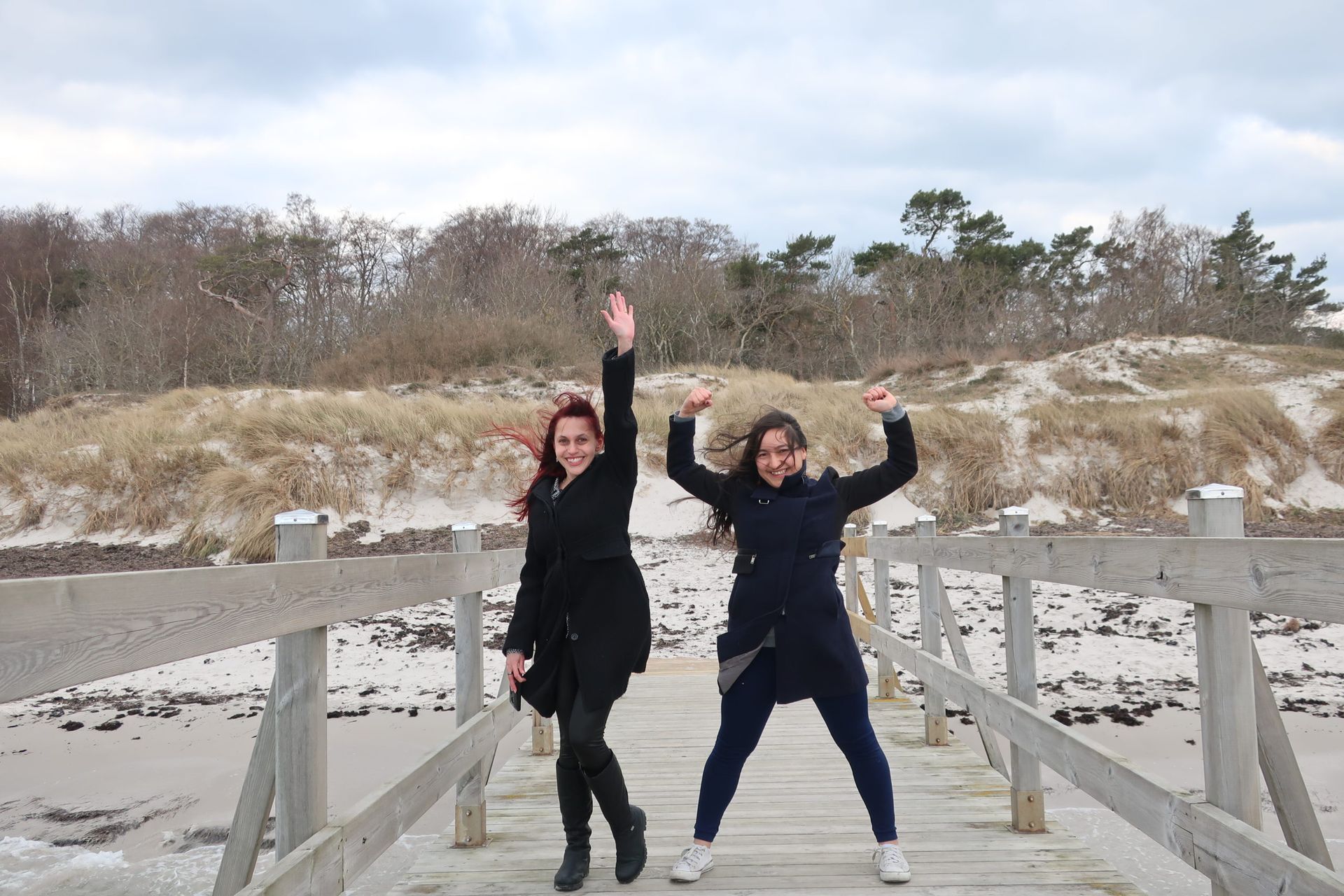 Two women cheering and jumping up and down on a jetty on a windy day. They are both wearing winter jackets.