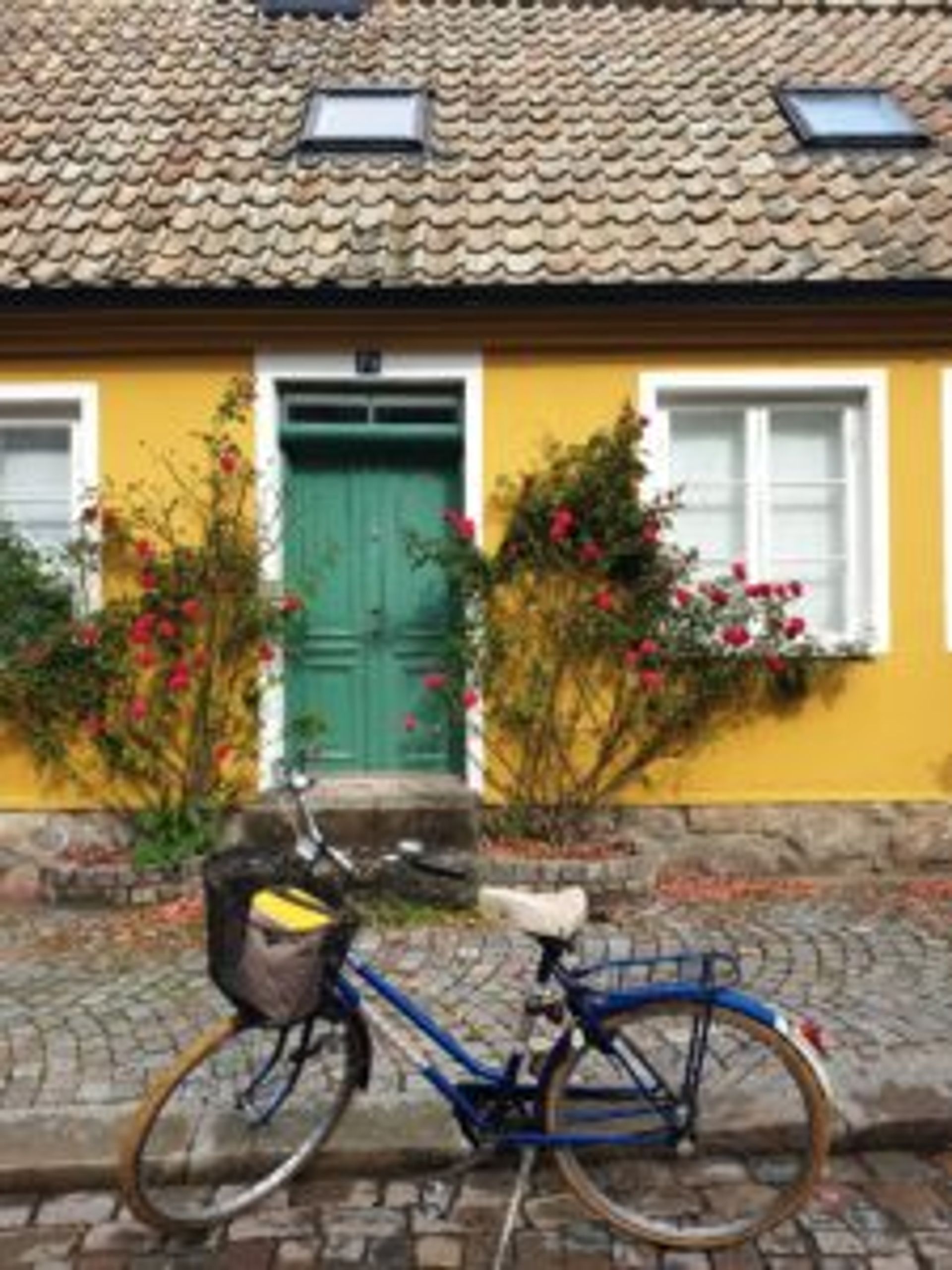 A blue bicycle is parked on a cobbled steet outside a small, yellow house.