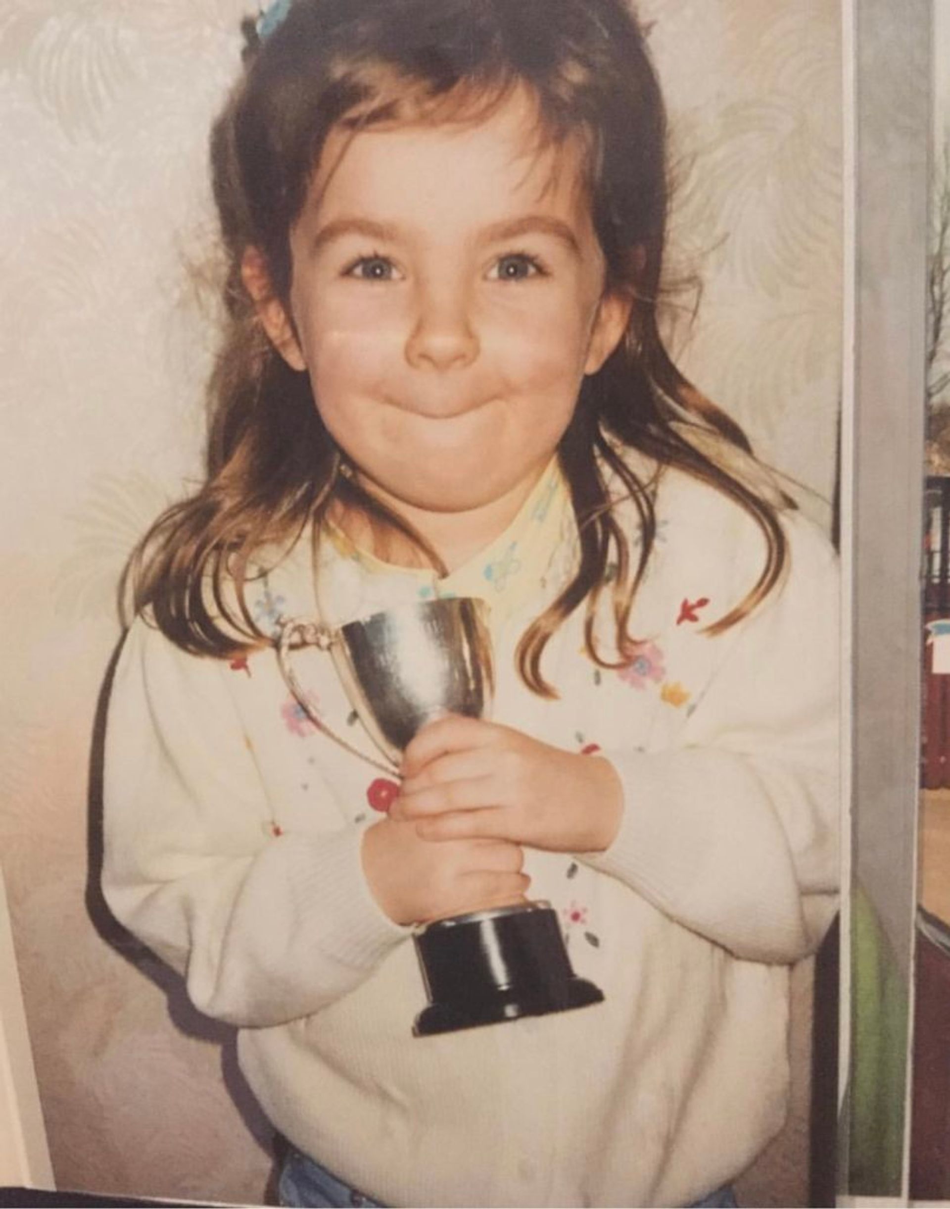 Emma as a child. She is wearing a white sweater and holding a little trophy. 