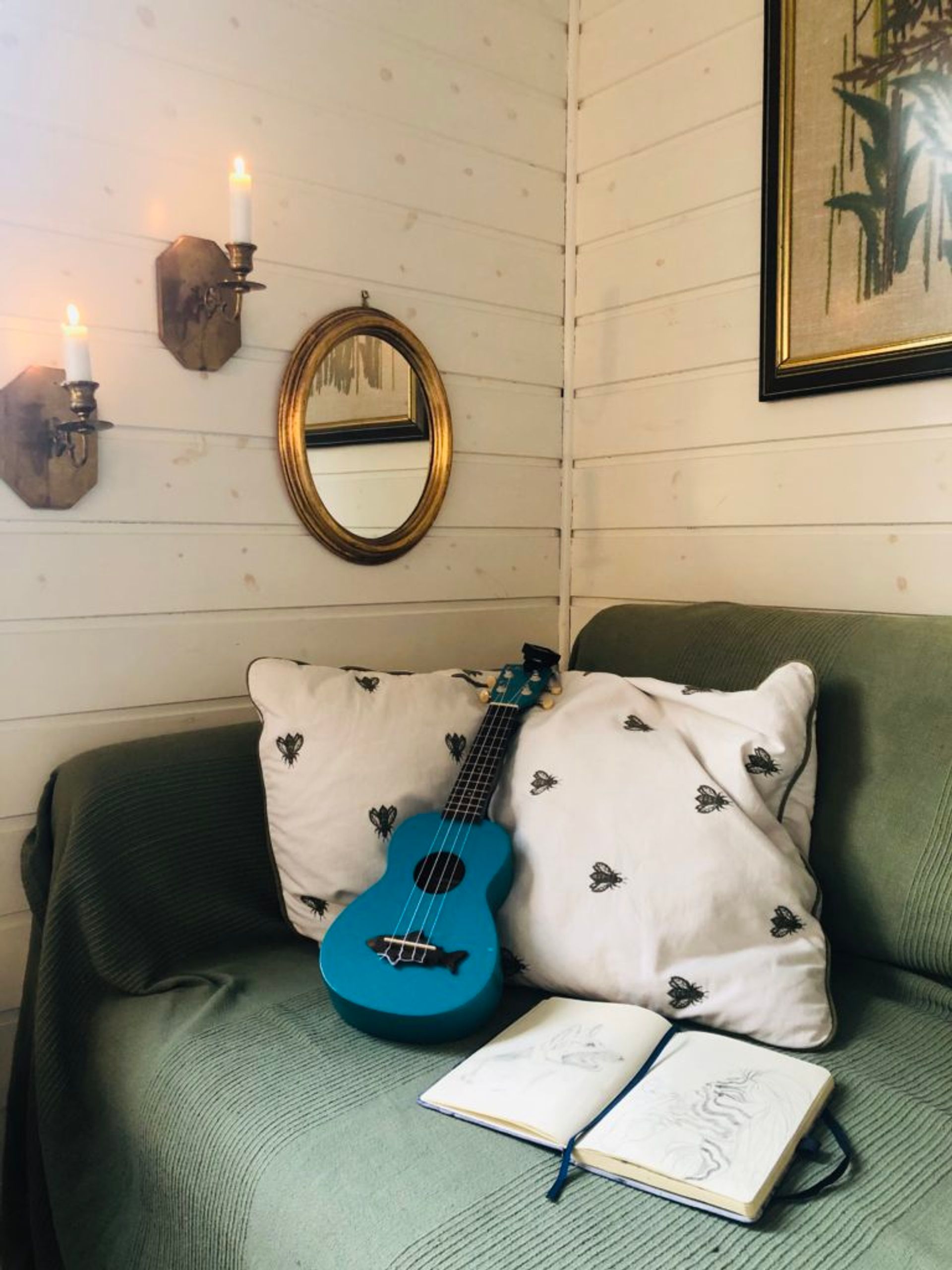 A sofa, covered in a green blanket. On the sofa are two cushions, a blue ukelele and a notebook.