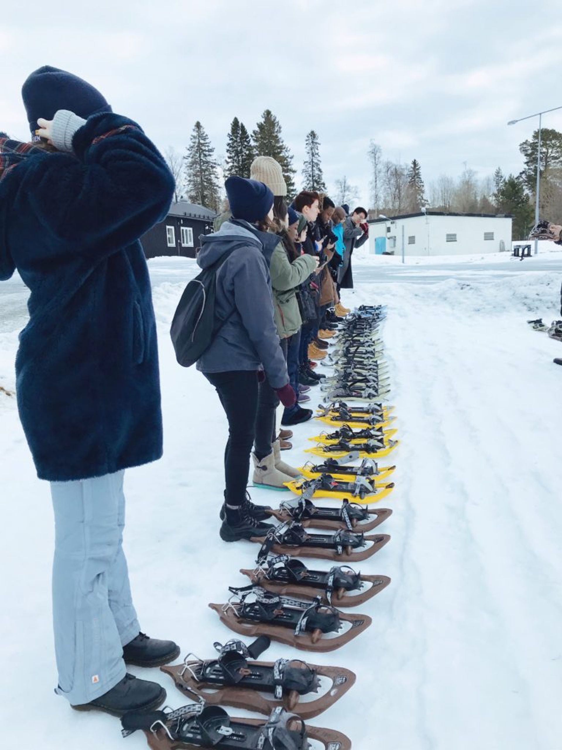 Students standing in a line with snowshoes set out in front of them. Some are taking photos of the snowshoes.