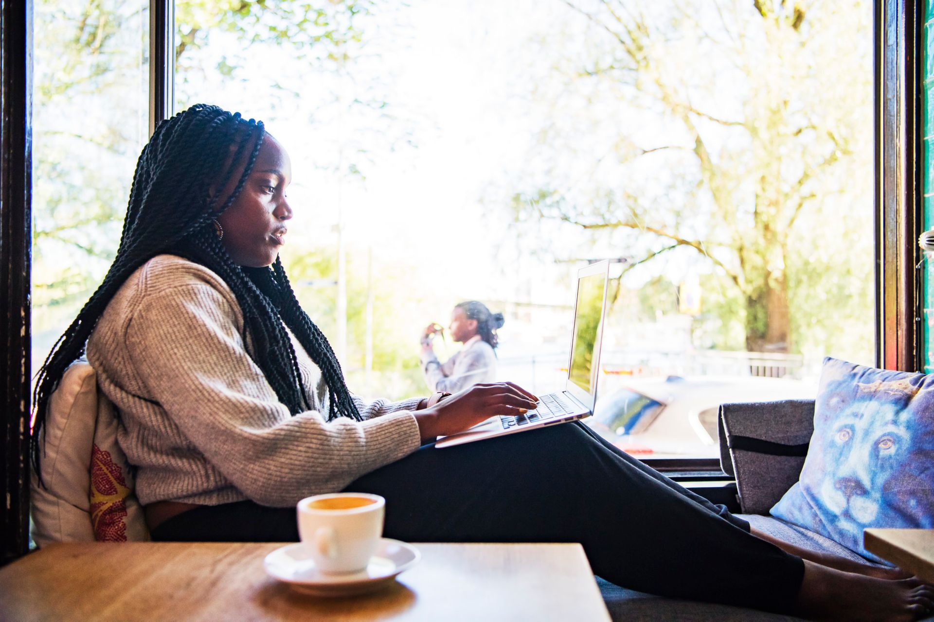 Woman sitting by a large window in a cafe, working on a laptop.