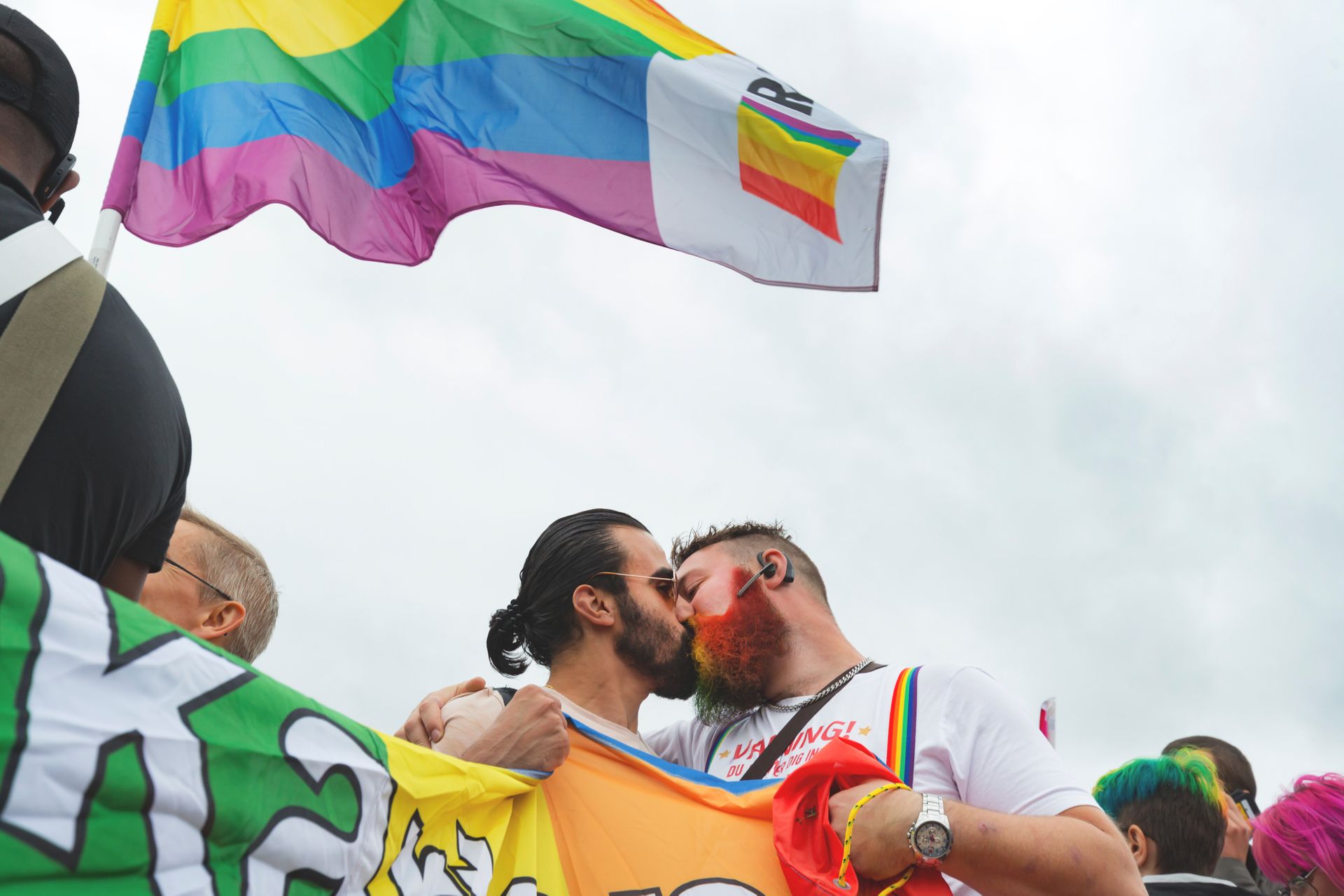 Two men kissing. They are holding a Pride flag and another Pride flag is waving beside them.