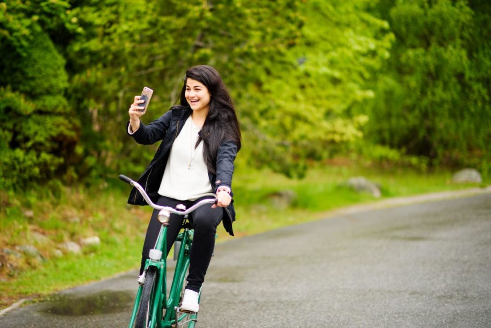 Marin cycles down a road on a green bicycle. She is holding a mobile phone in her right-hand.