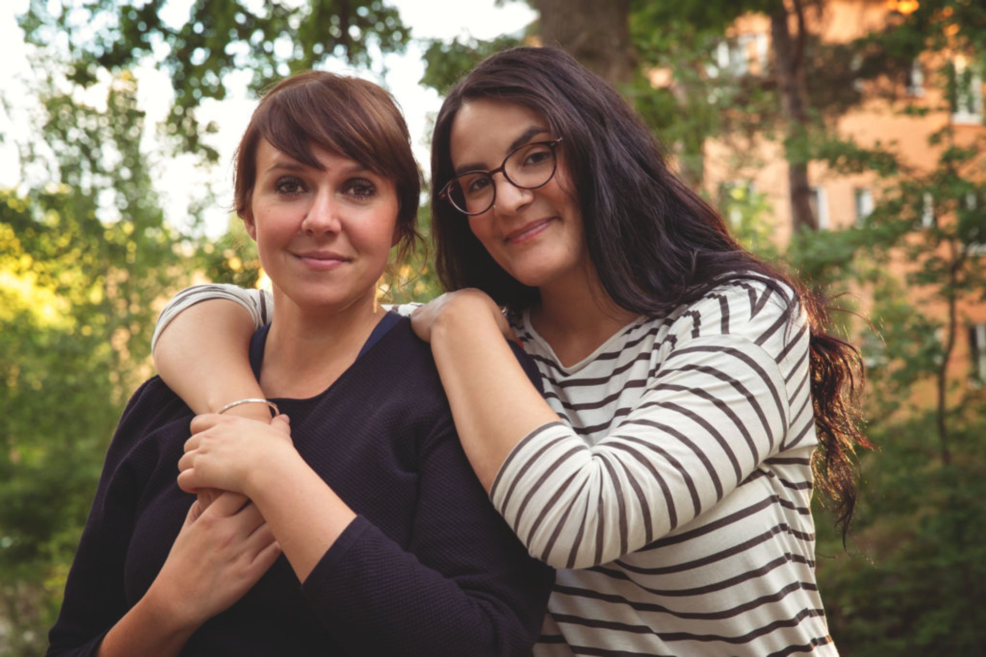 A couple stands outisde together. One woman has her arms around her girlfriend's shoulders.