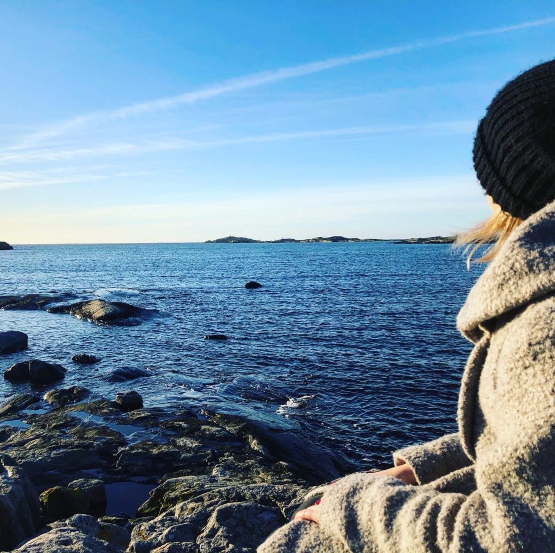 Sara, dressed in a winter coat and hat, sits on a rock by the sea staring out at the view.