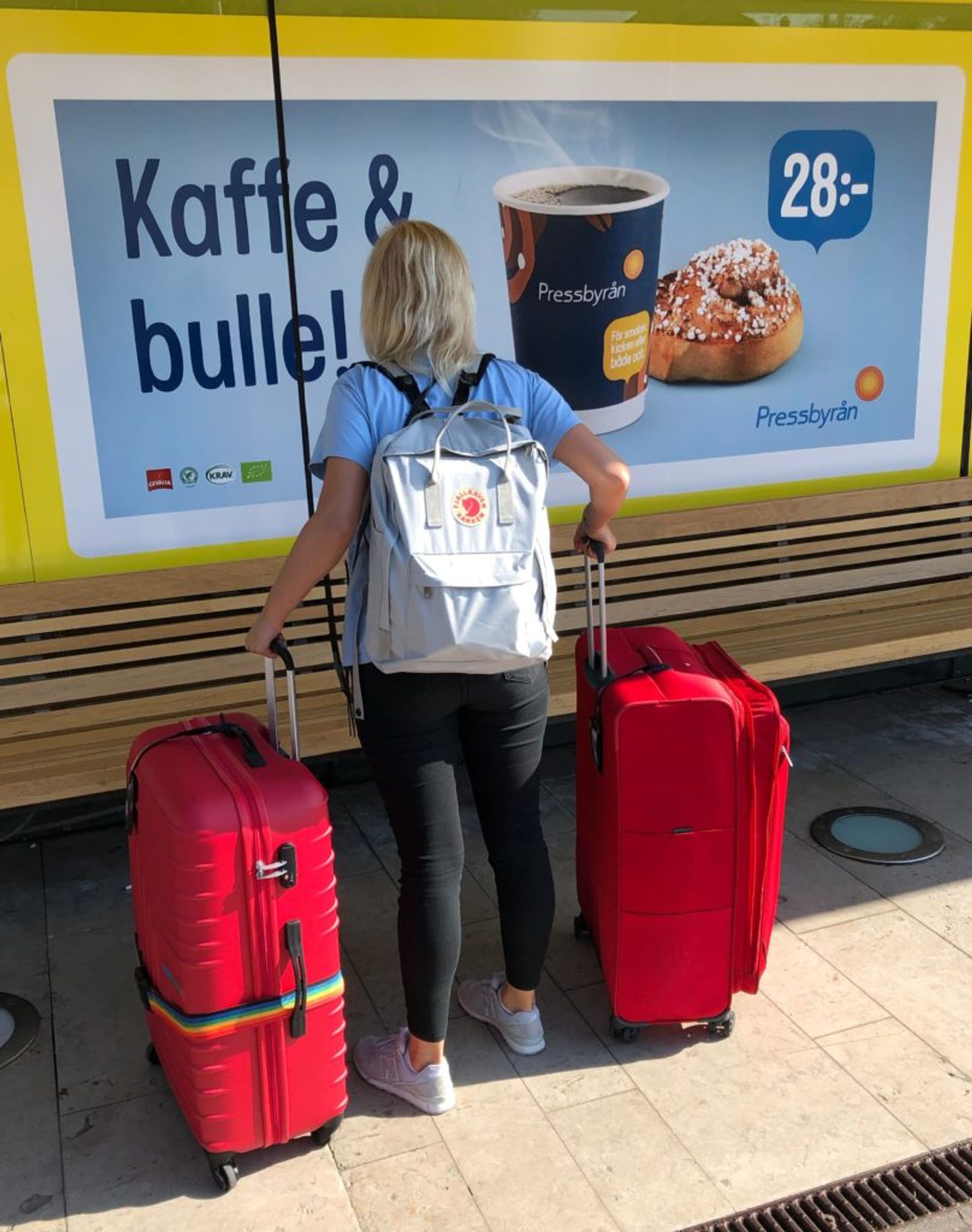 Sara stands in front of a shop at the airport in Sweden holding two large red suitcases.