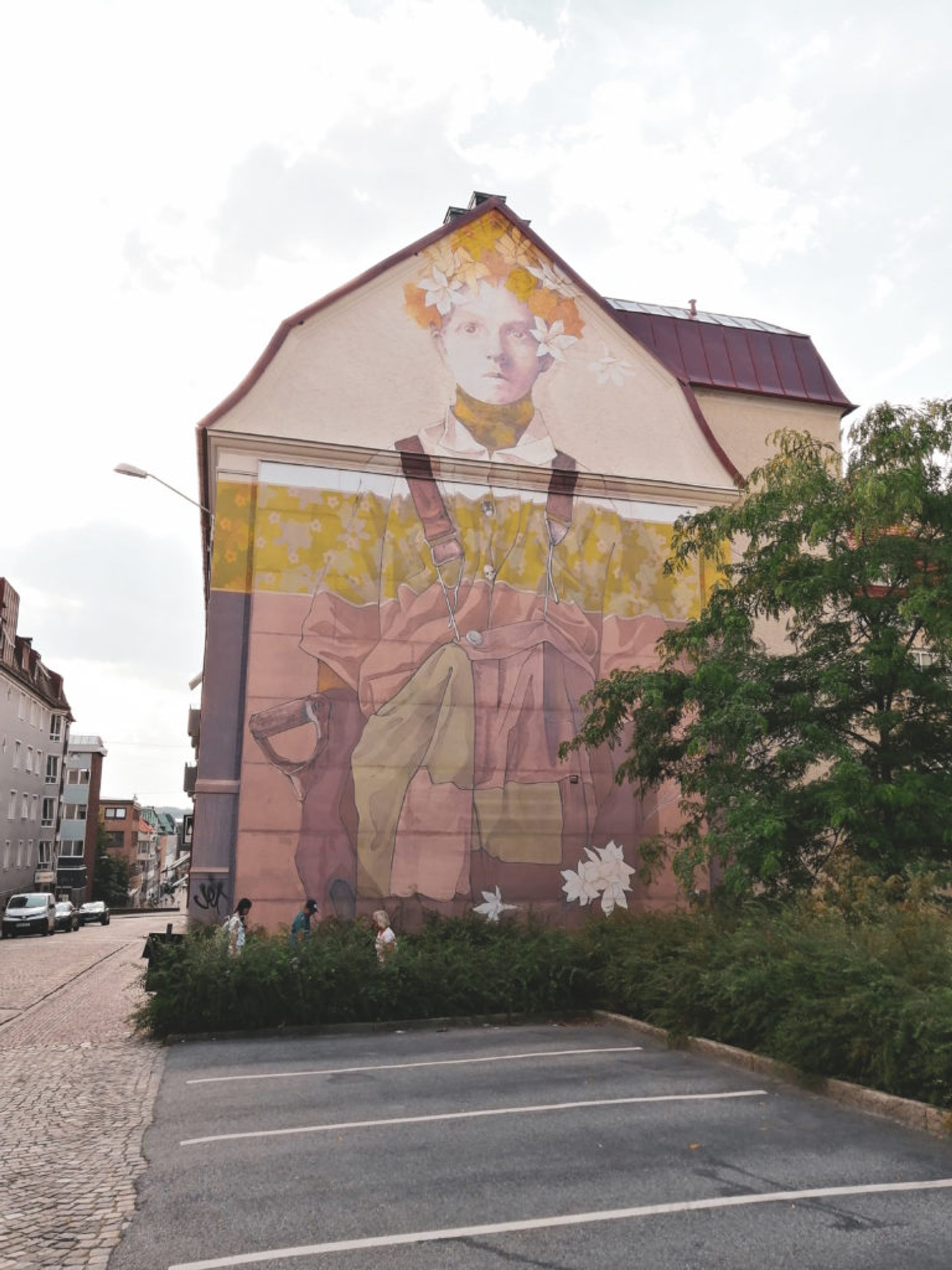 Washed out mural of person wearing dungarees and flower crown on the side of a residential building.