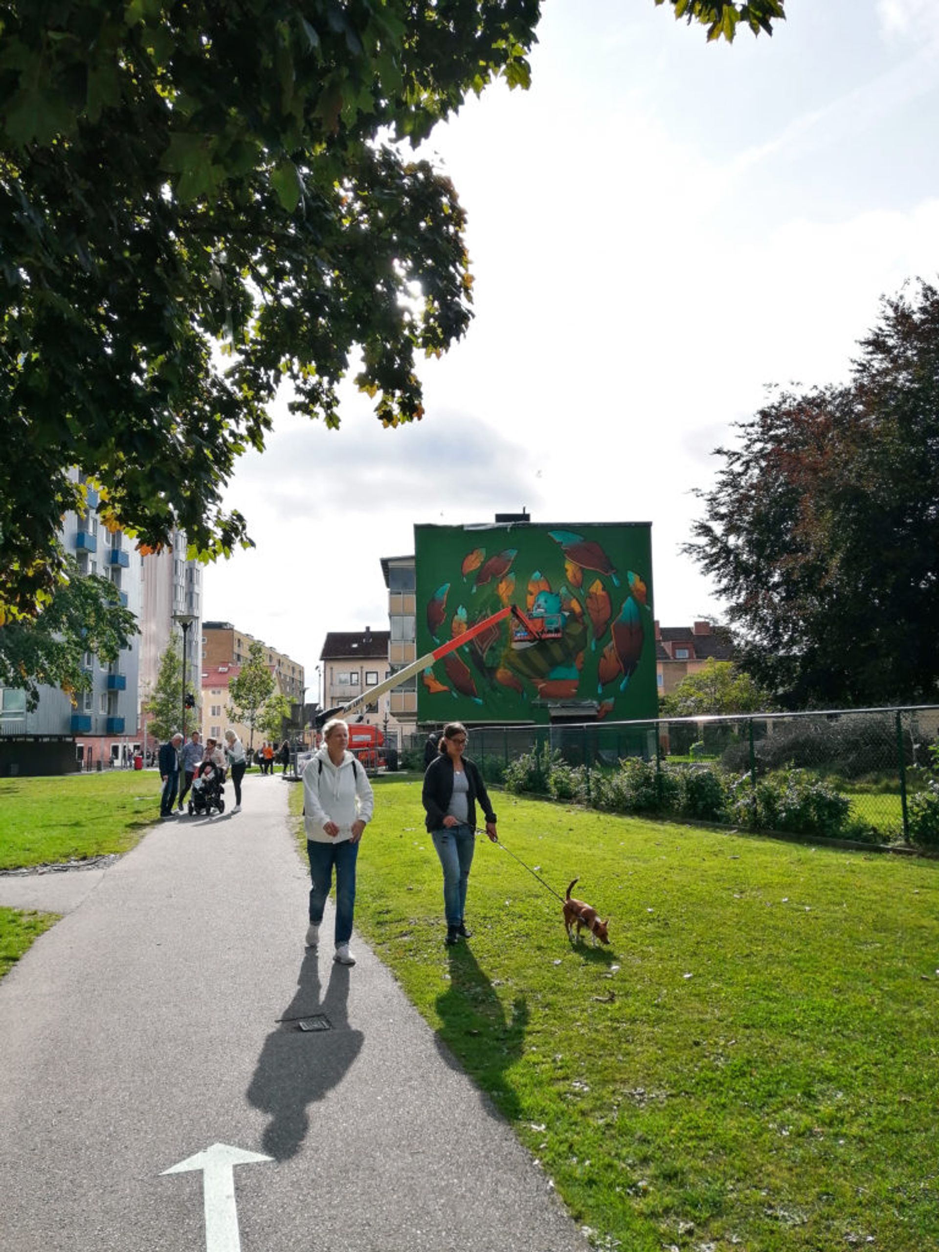 Women walking a dog in front of a mural being painted in Norrby.