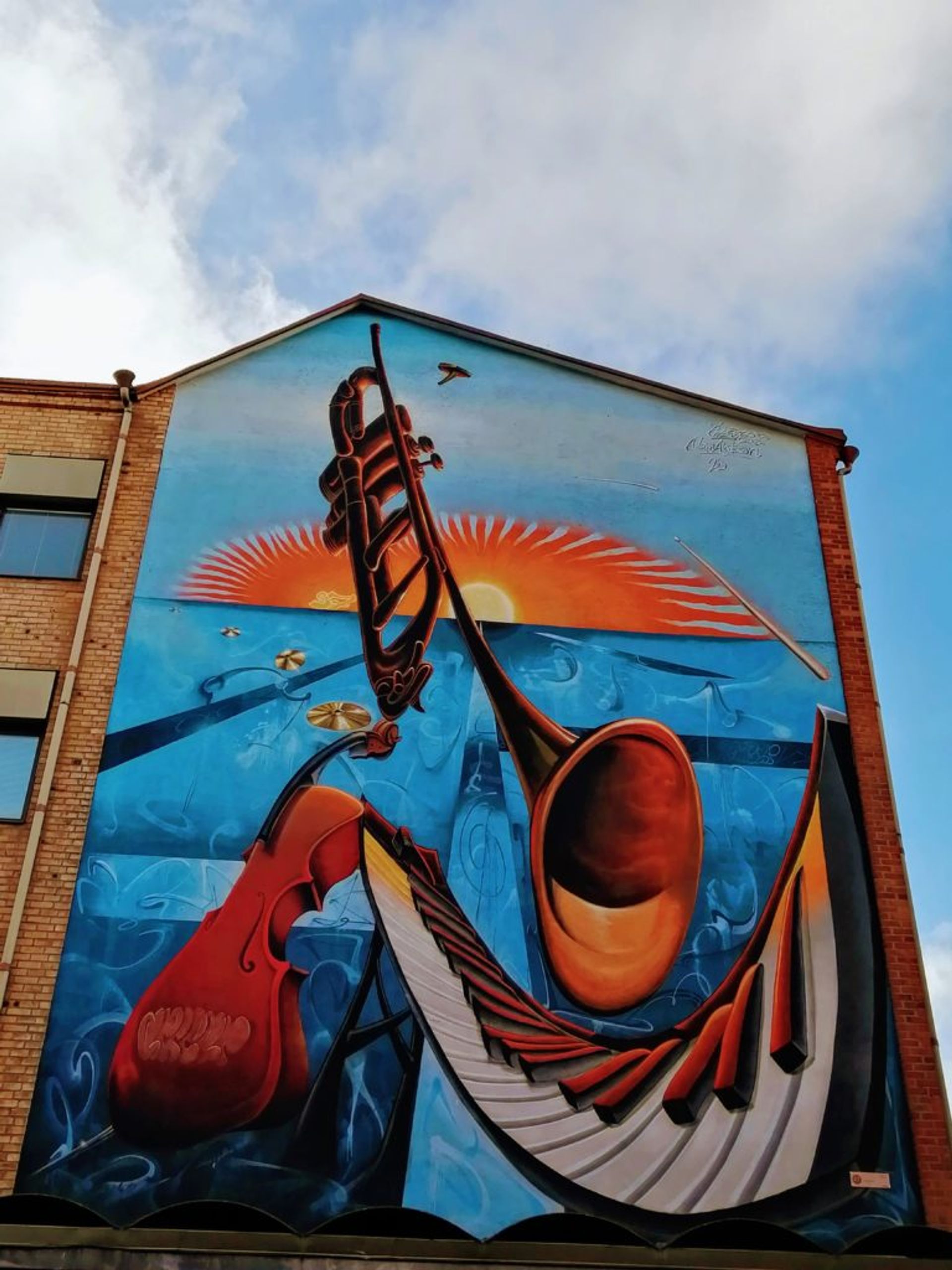 Bright blue and orange mural of musical instruments at sunset on the side of a red-brick building.