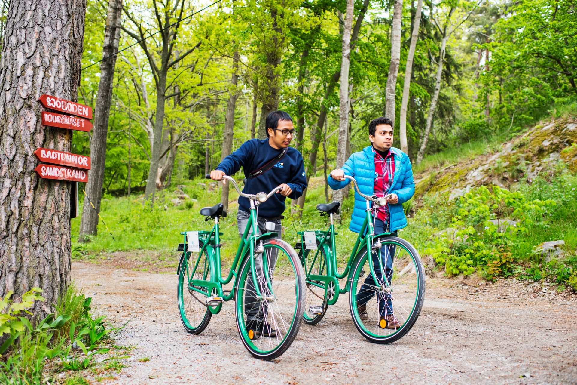 Two international students walking with green bikes in a forest.