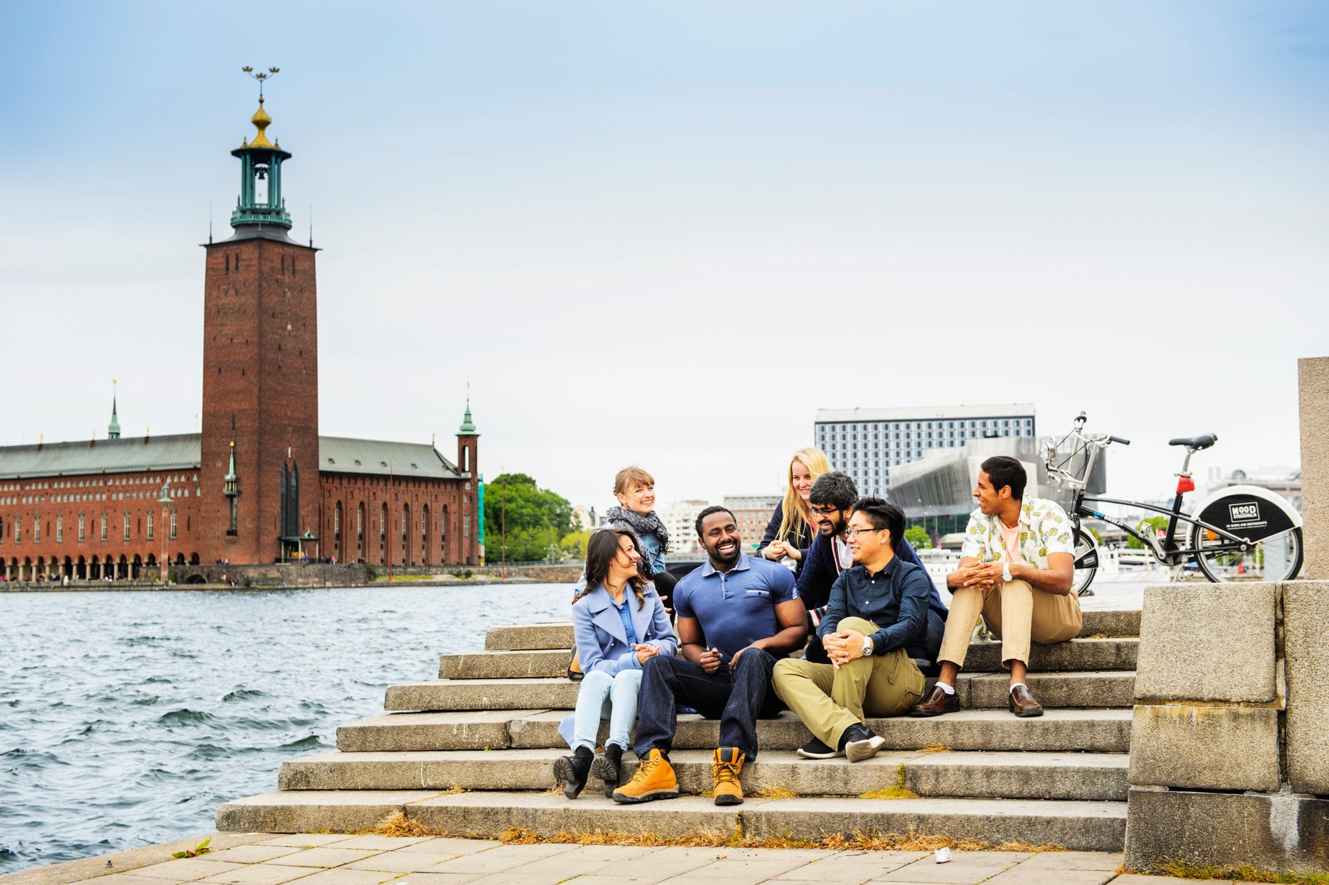 A group of students sitting on stairs, with the Stockholm City Hall in the background.