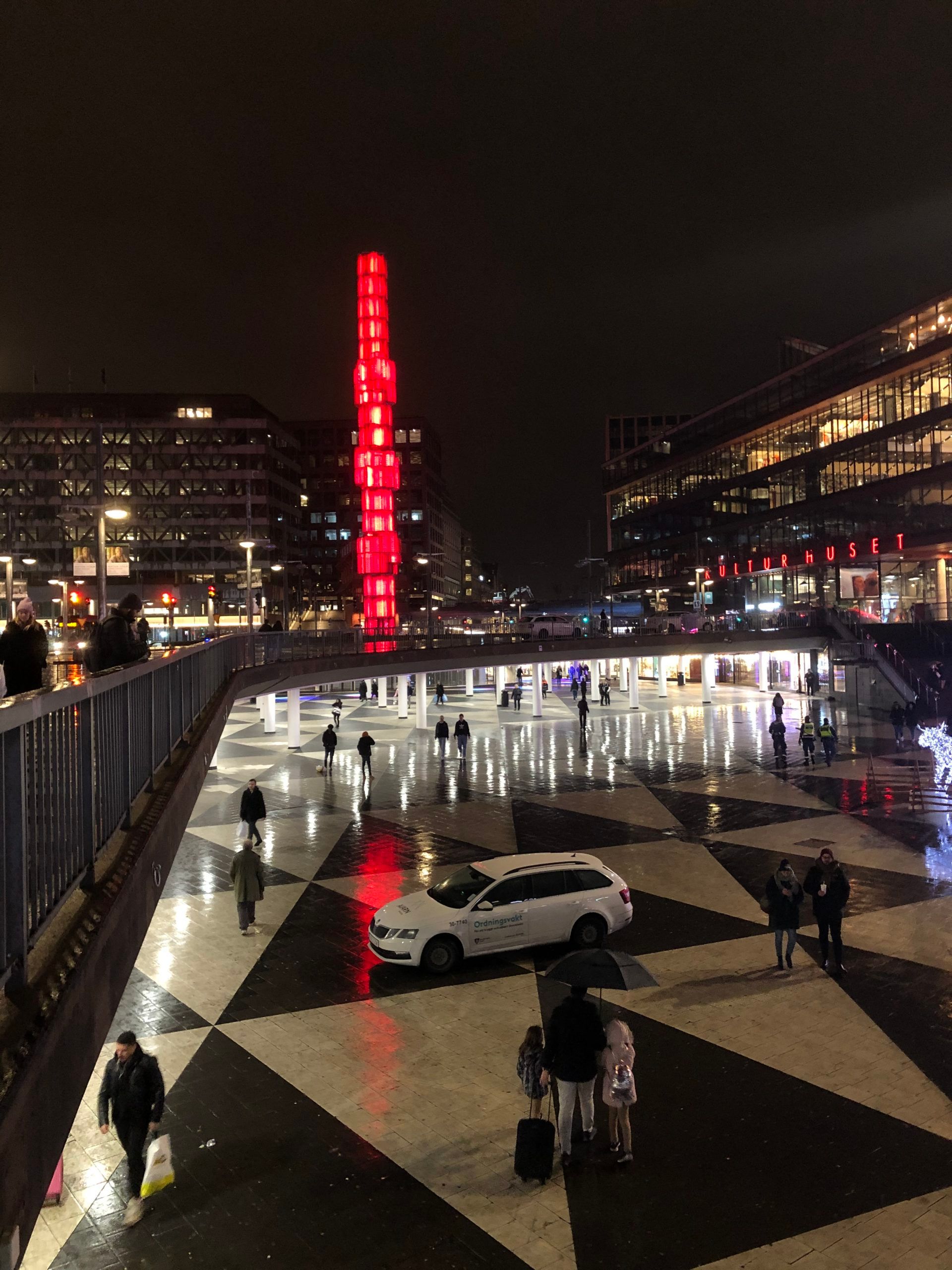 A tall tower building lit fully in red lights onto a shiny, black and white triangle designed pavement, behind a bridge reflecting scattered lights. Pedestrians walking under the bridge and a car parked in front.