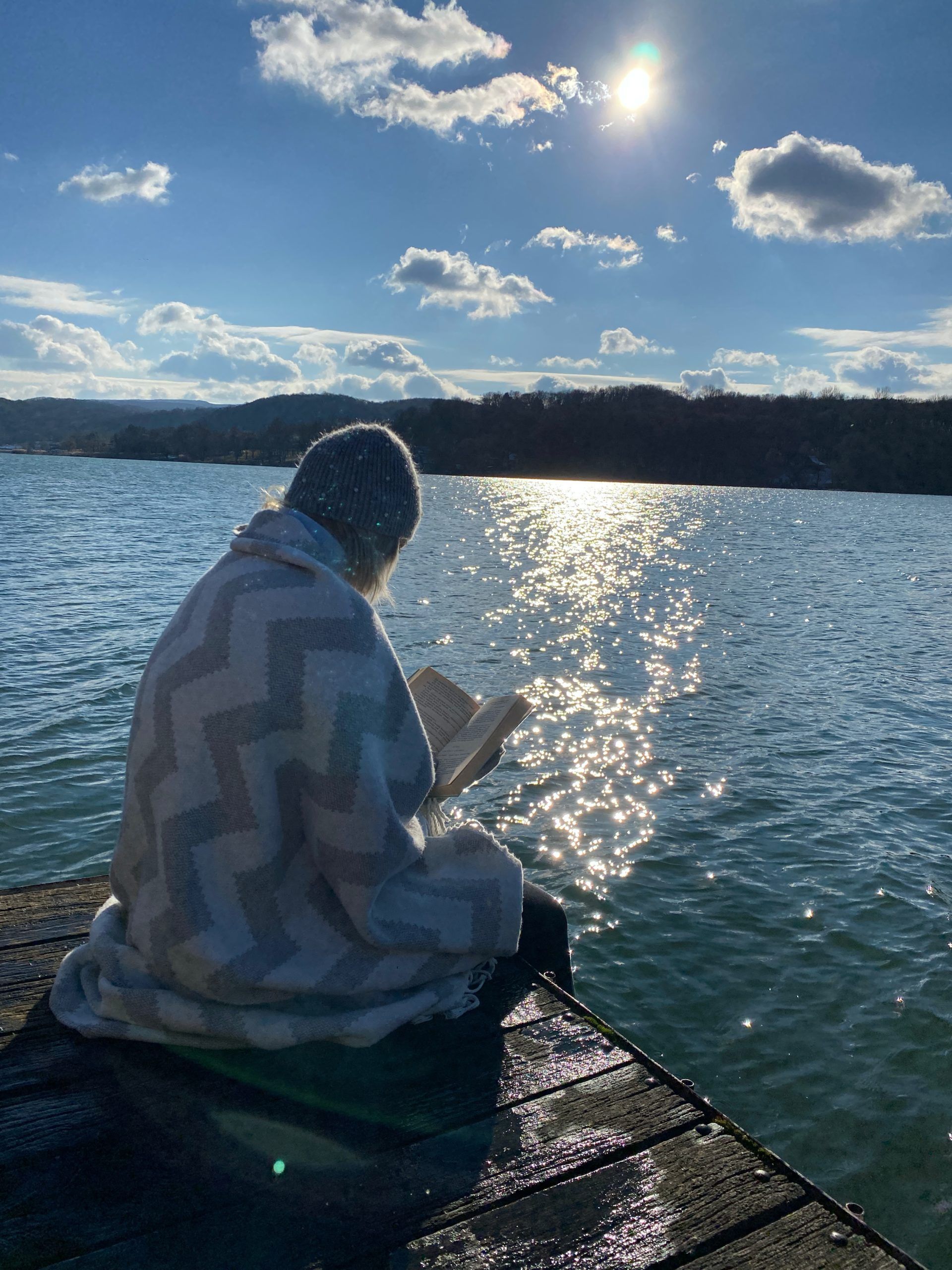 Picture of me, wrapped in a blanket, with a hat on, while reading on a pier at a Swedish lake 