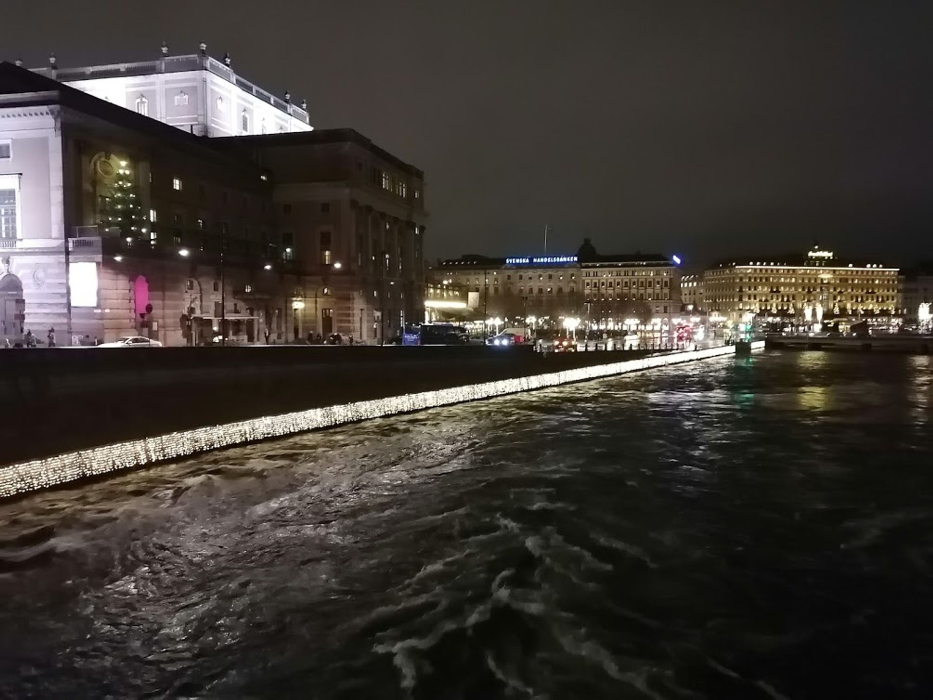 Large historical building with bright lights facing a canal bank, with fairy lights along the canal border