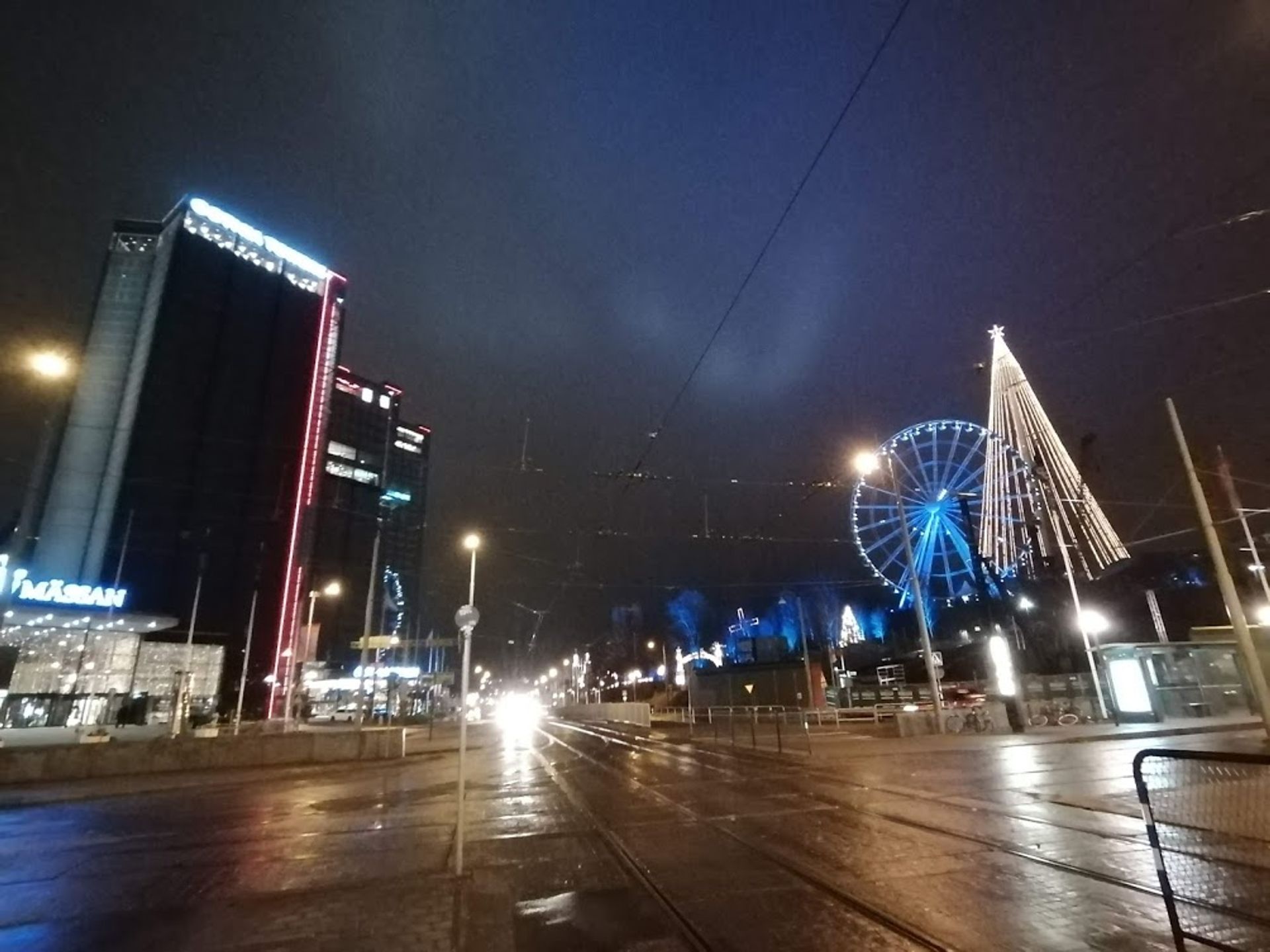 lights reflecting on a glistening street, with a lit up ferris wheel and tree light scuplture in the background