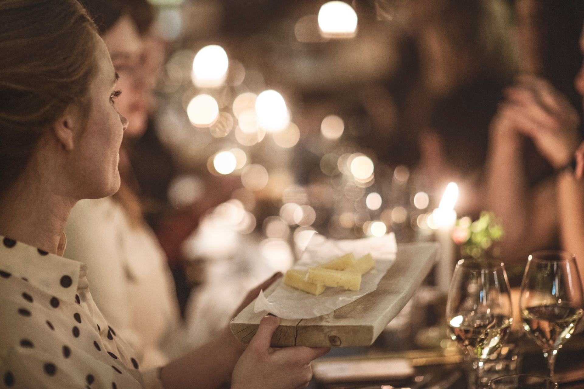 A woman holding a cheese board and in the blurry background you see people and lights