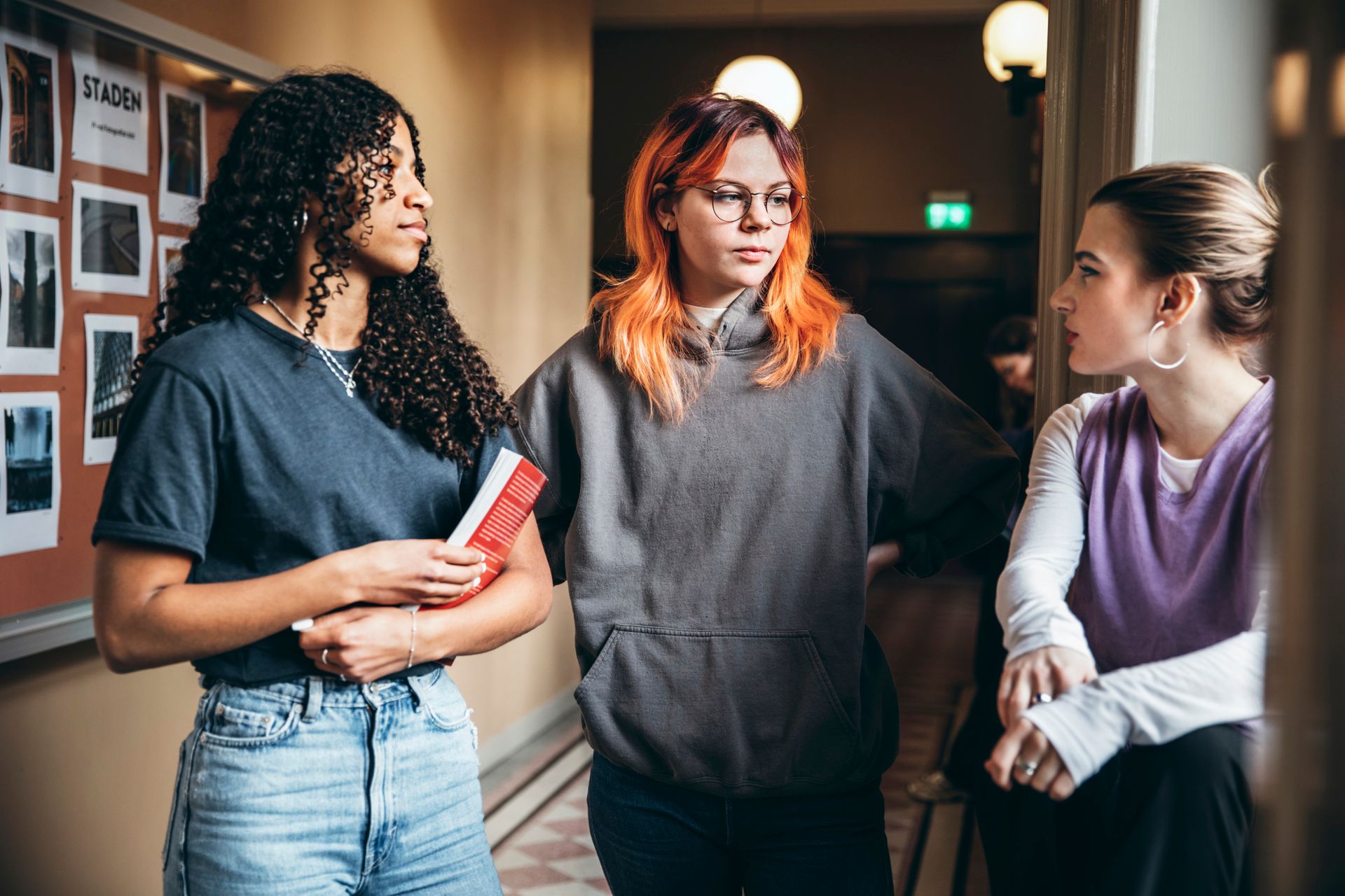 Three teenage girls stand in a hallway, talking.