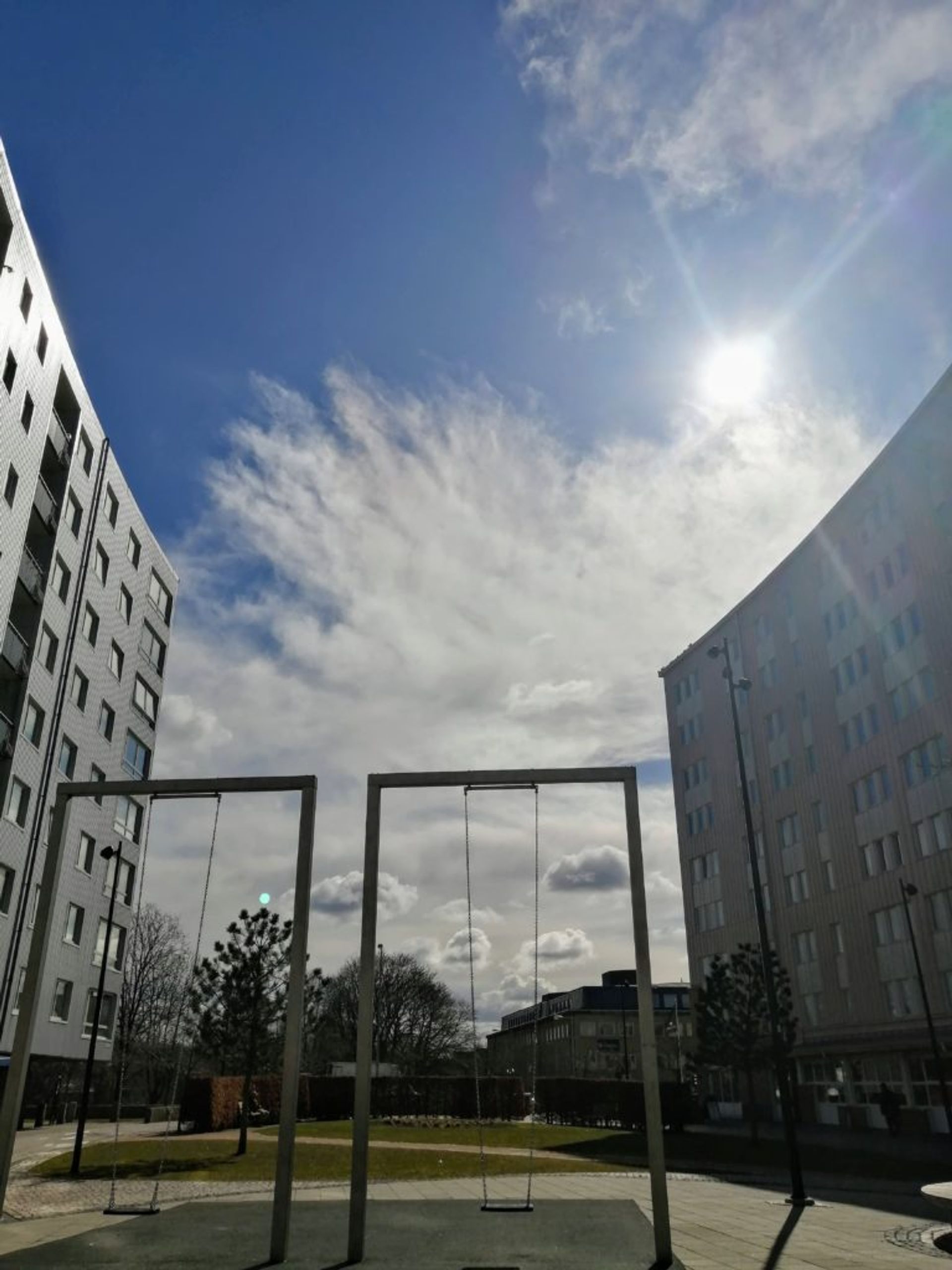 A swing set in between two tall apartment buildings with clouds overhead passing to reveal the sun