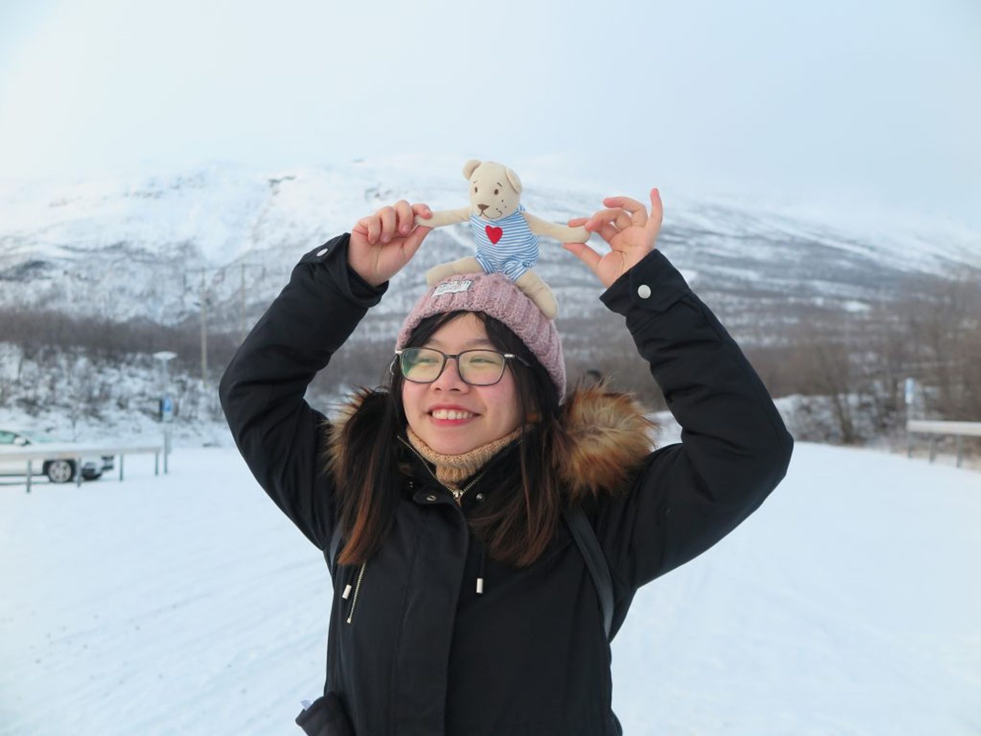 Young girl holding a teddy bear upon her head and smile happily with snowy mountain in the background 