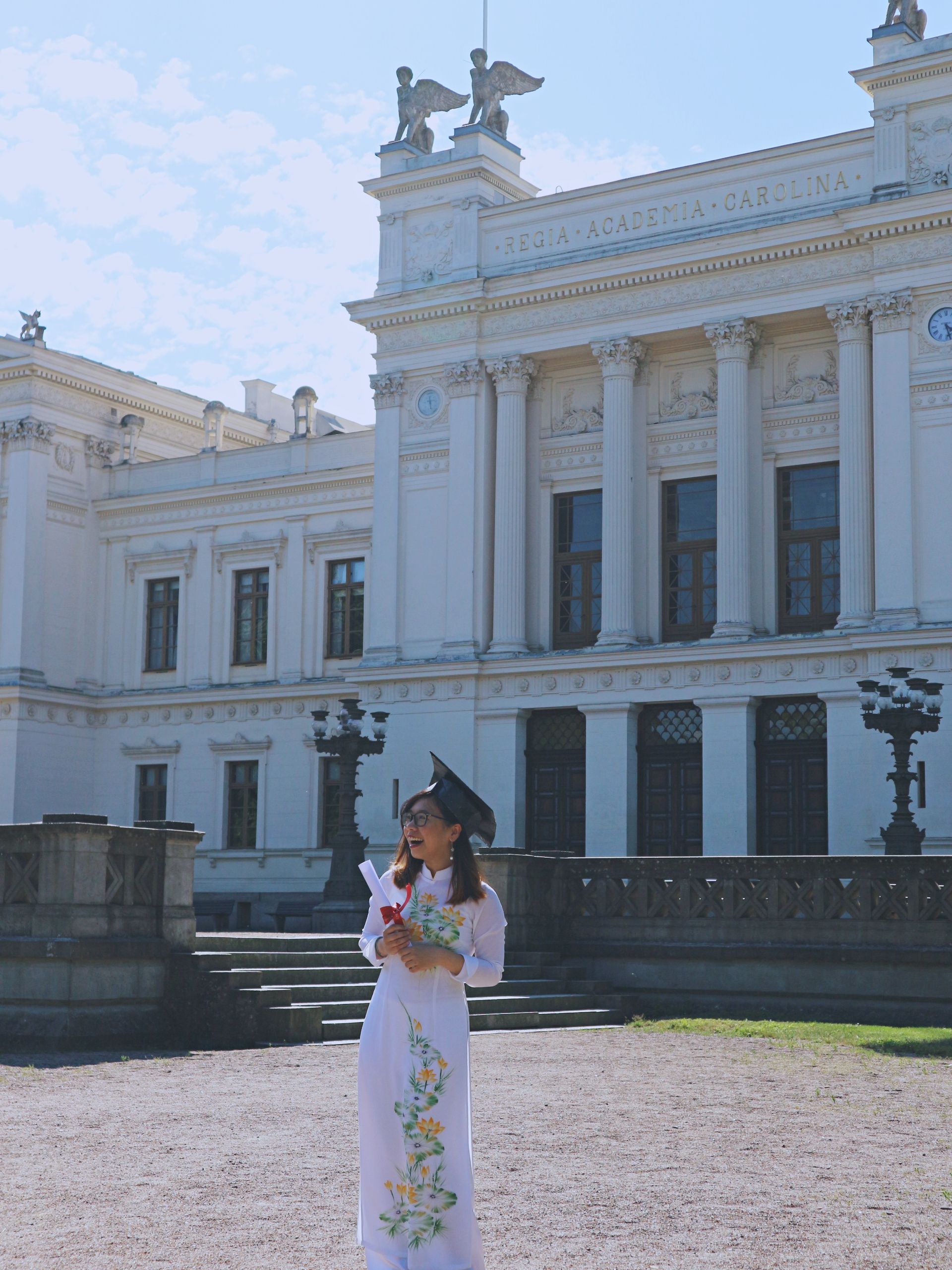 A young girl wearing traditional Vietnamese dress and holding a degree in her hand in front of the Lund University main building