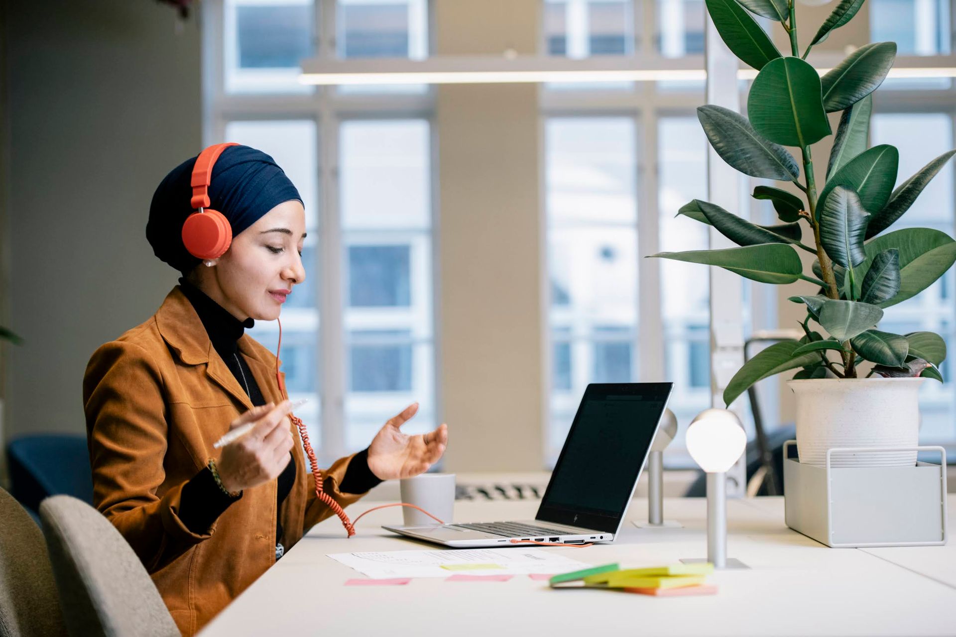 A young woman wearing headphones over a hijab, sitting a a desk and working in front of a laptop