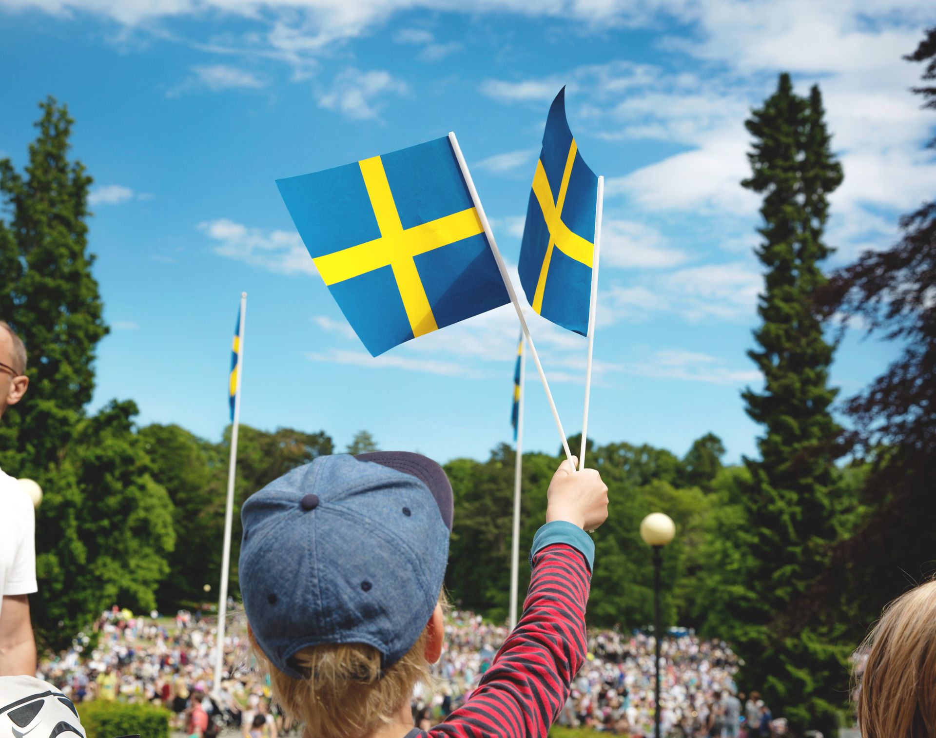 A child in the foreground is waving a Swedish paper flag of blue and yellow. In the background there is a crowd of people in a field surrounded by trees.
