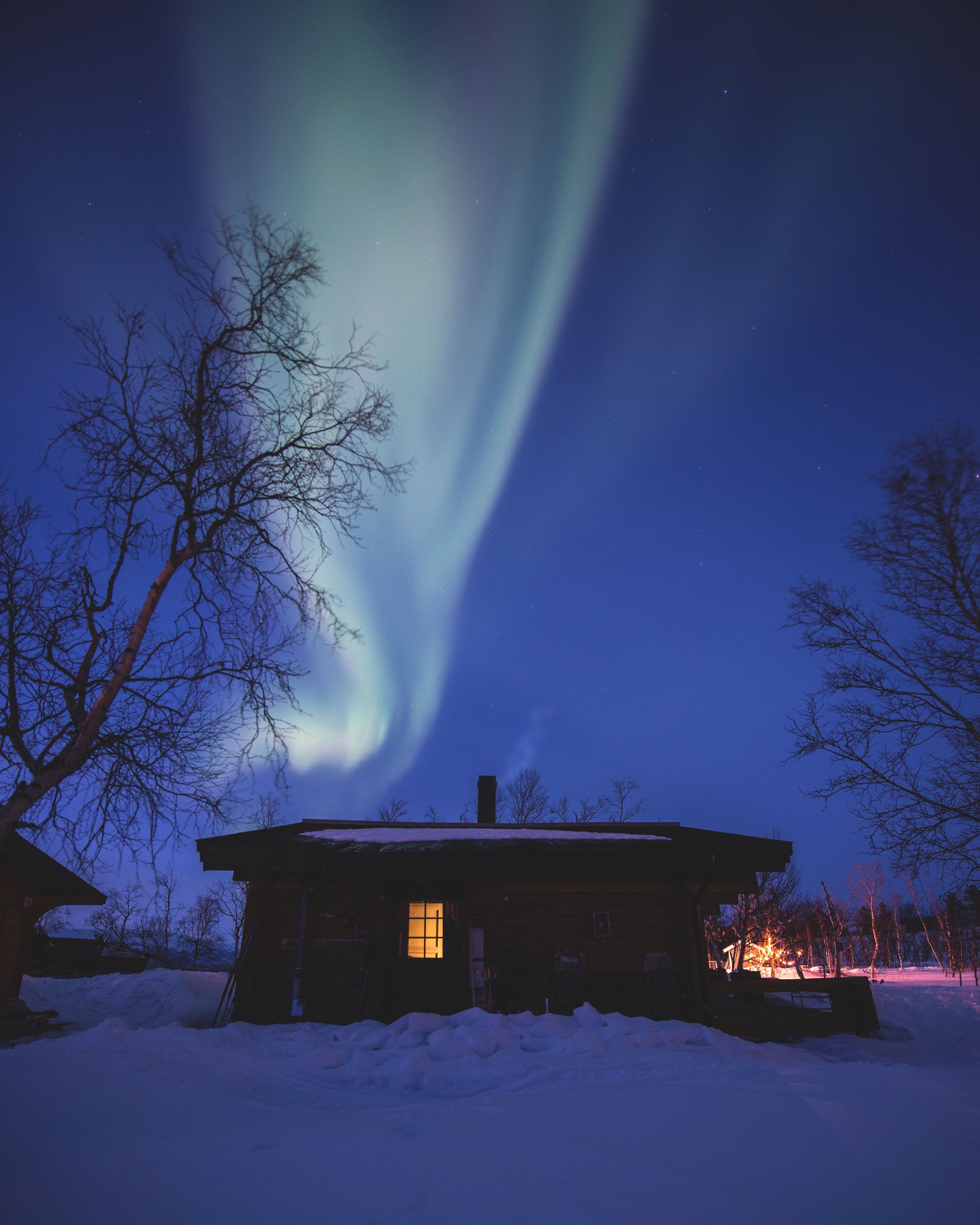 The Northern lights is seen above a mountain cabin. It’s winter and snow on the ground. It’s night and light is on in the cabin.