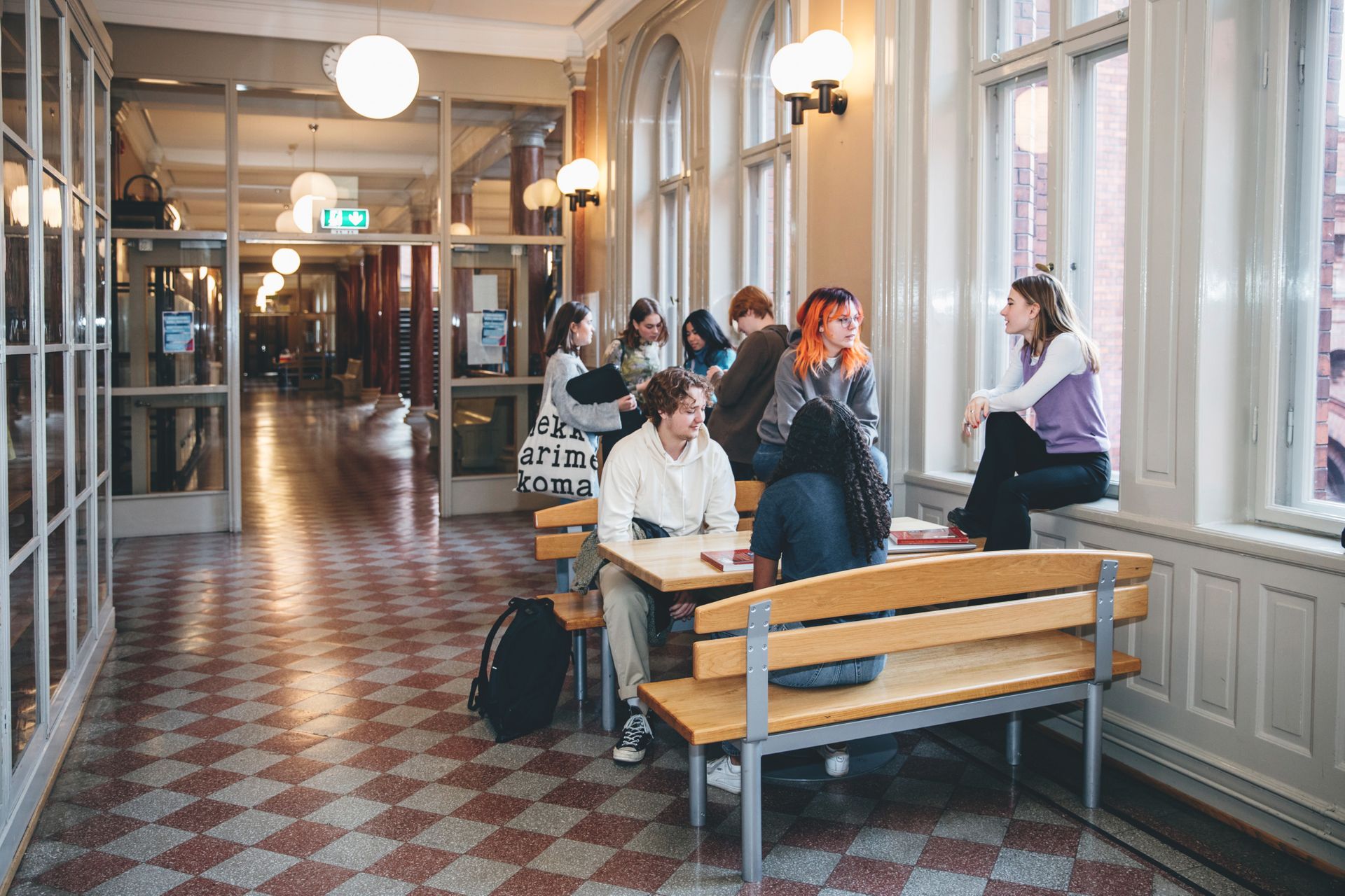 Teenagers sit together by tables in a long hallway.