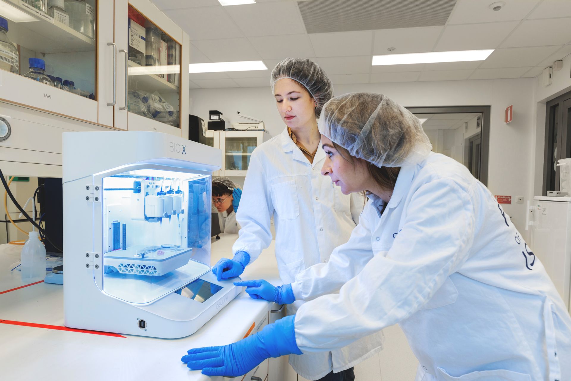 Two women wearing hairnets stand in front of a bioink 3D printer.