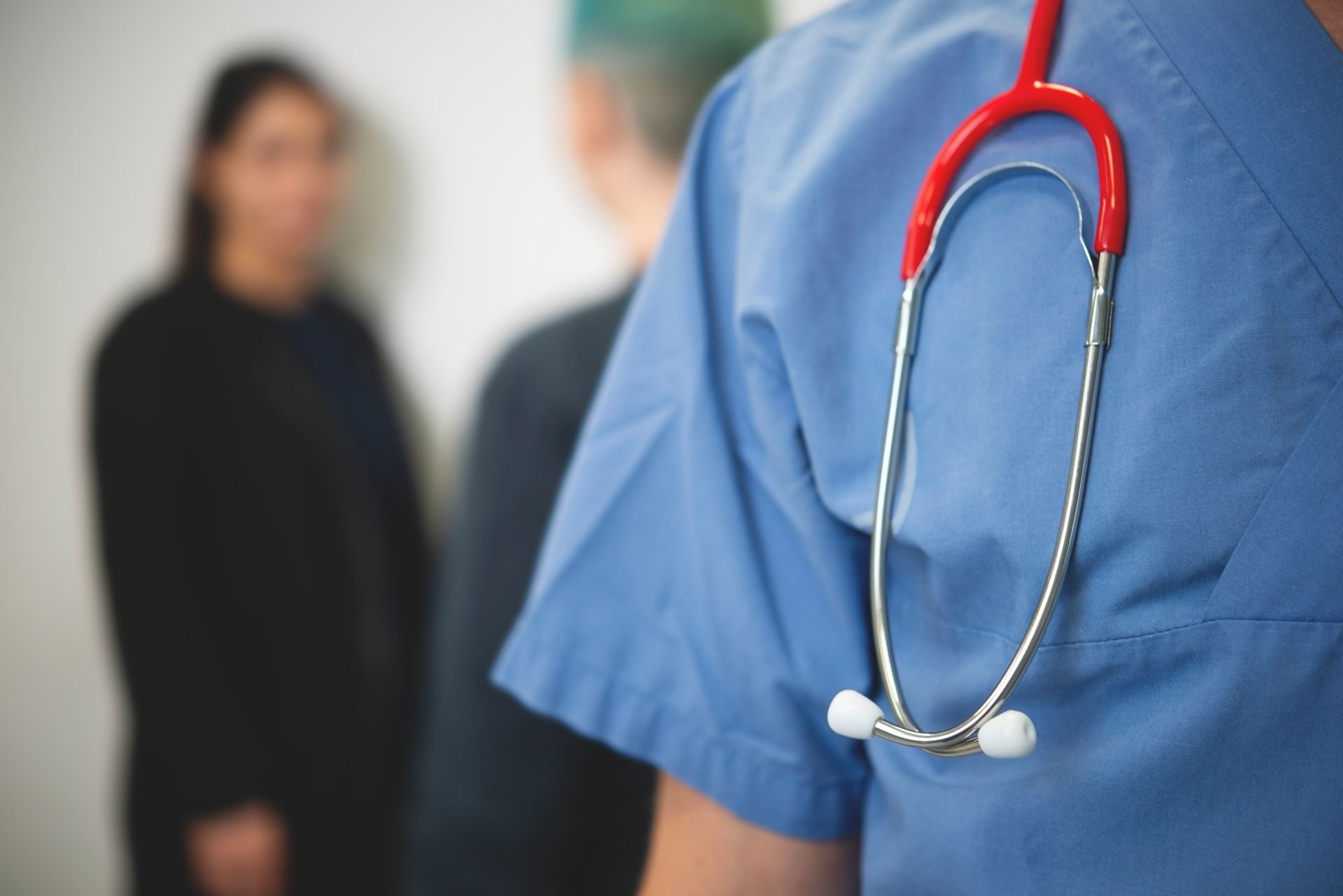 A stethoscope is slung over a shoulder in the foreground. Two people face each other in the background.