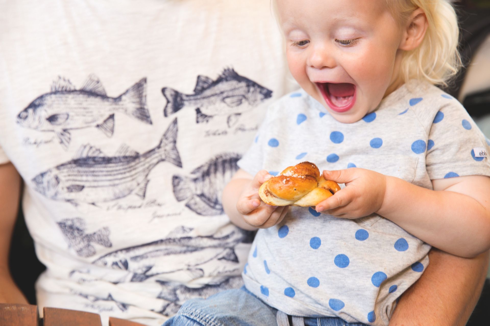 A happy child with an open mouth holding a cinnamon bun sits in a lap.