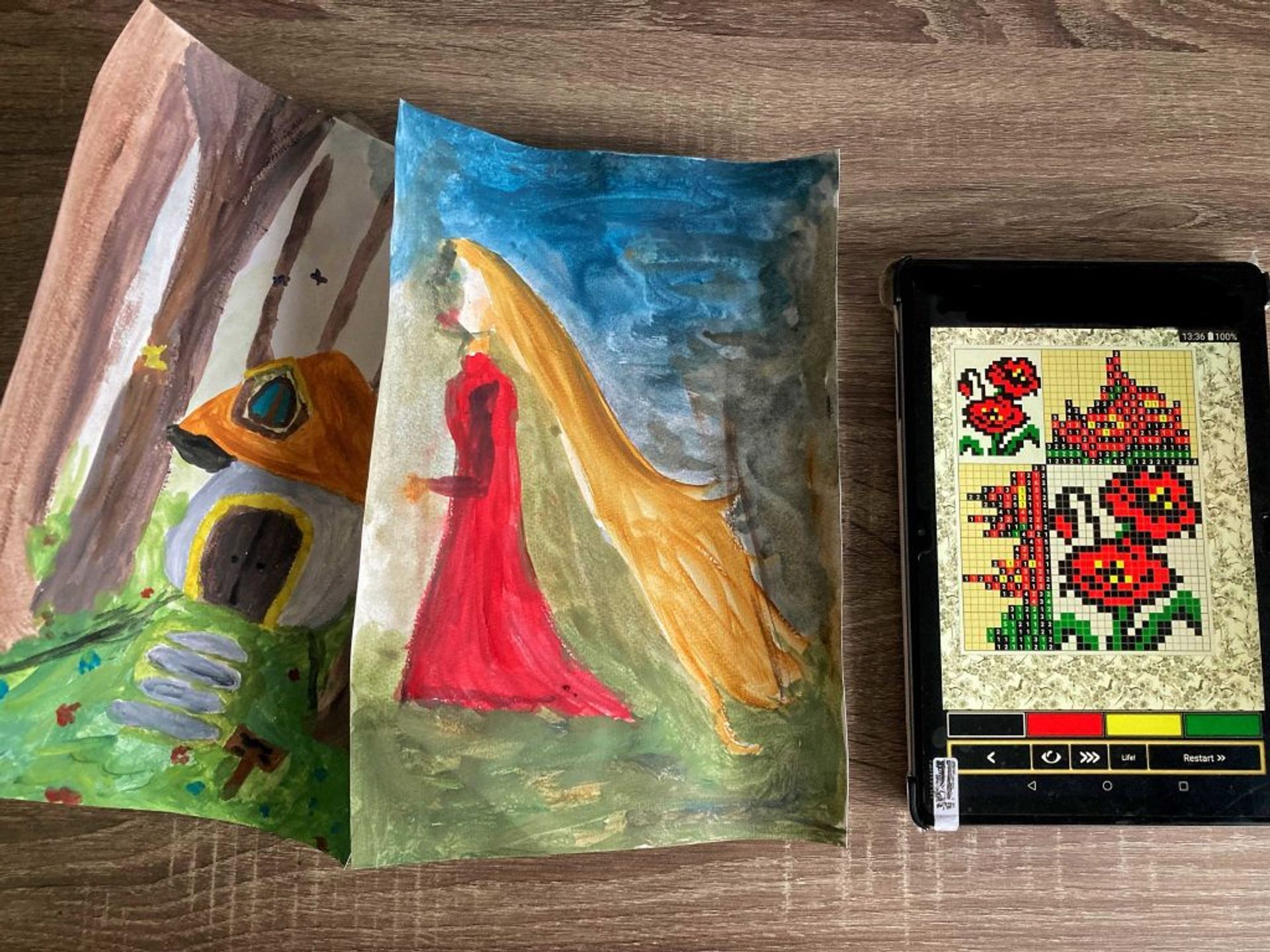 Two paintings (one of a small cottage in the woods and another one of a girl with long blonde hair) and a tablet with a nonogram. 