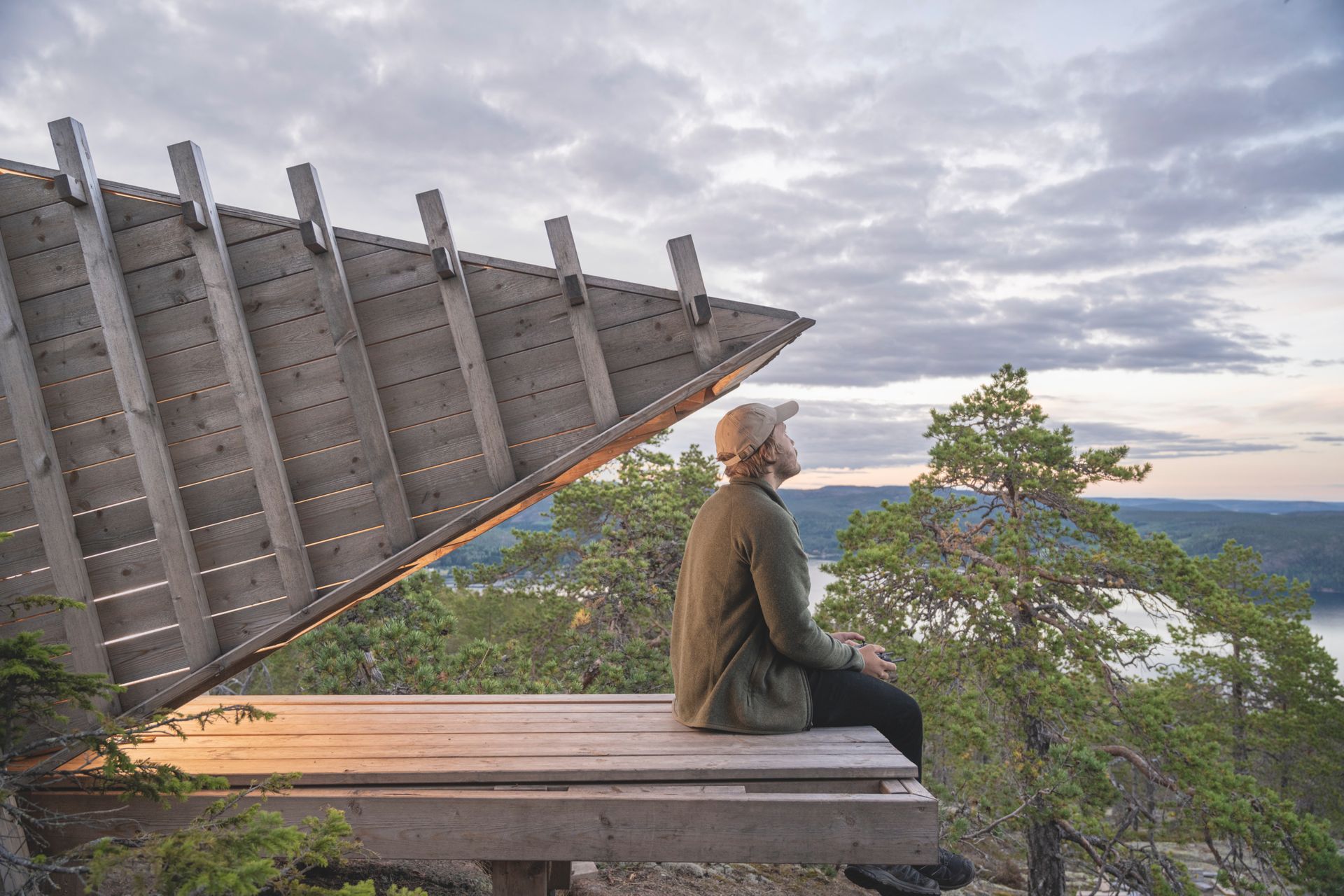 A man sits outside a minimalistic and modern style hut or shack, looking out over the nature, forest and lake of the Swedish High Coast.