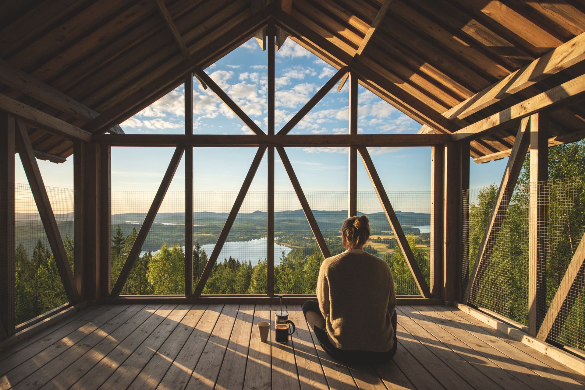 A woman sits on the floor of an open loft in the morning light, with freshly brewed coffee at her side, overlooking the mountains and the forest in Hälsingland.