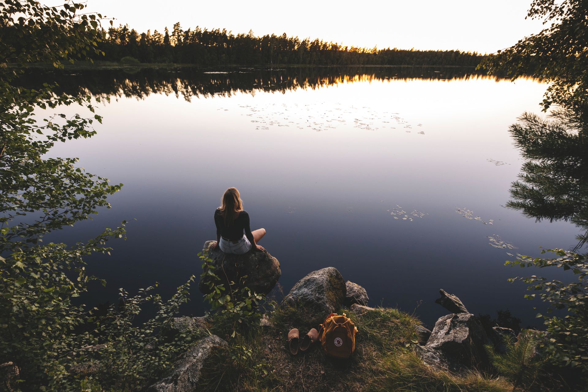 A woman sitting on a small rock looking out over a calm lake, the surrounding forest is mirrored in the lake.