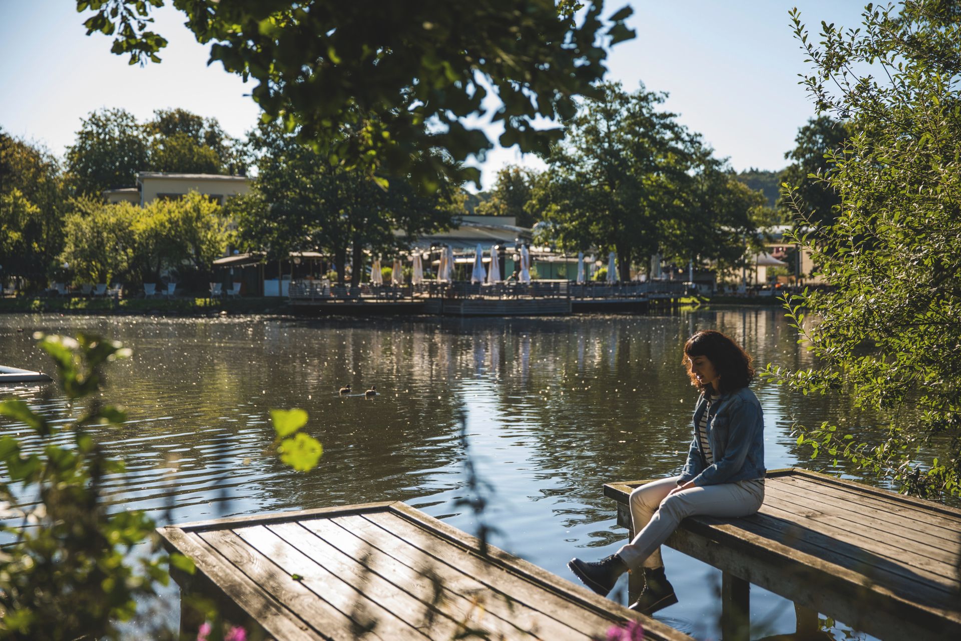 A woman sitting on a jetty next to a pond surrounded by greenery in a park. In the background you see an outdoor restaurant.