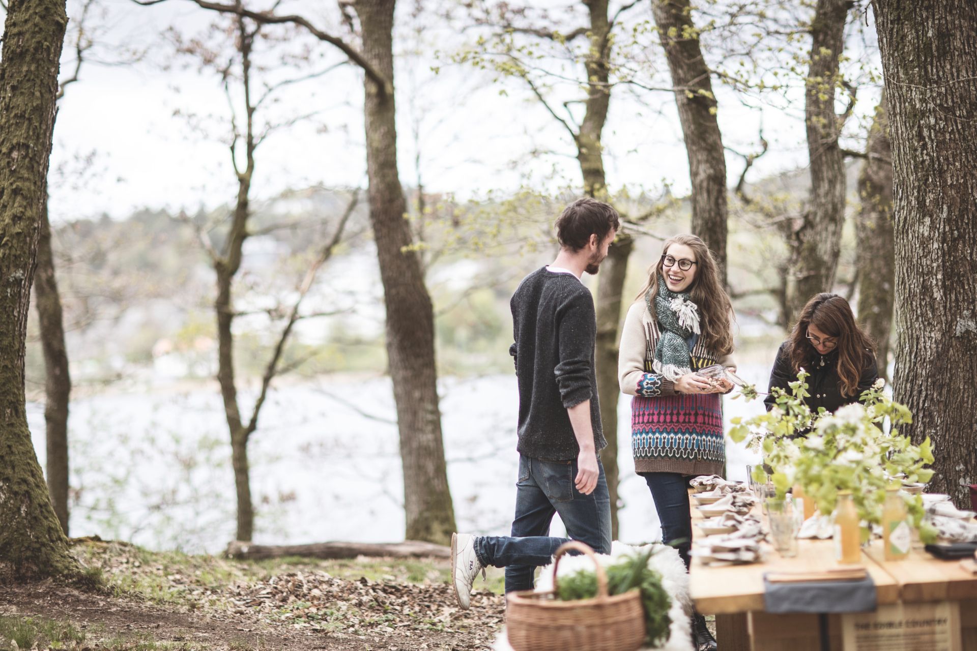 A wooden table set for dinner in a forest. Three people standing by the end of the table talking to each other.